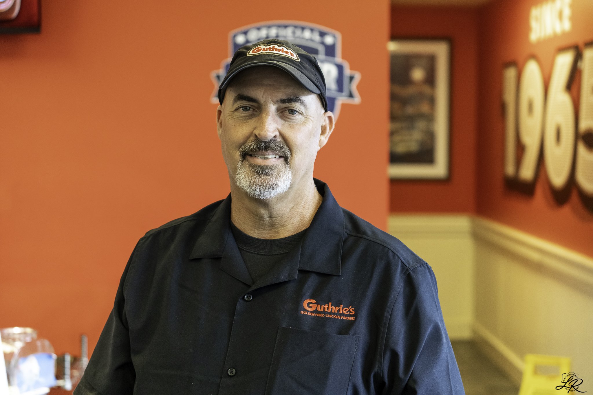 A man with a beard and mustache wearing a black hat and black shirt with a red logo that reads 'Guthrie's Golden Fried Chicken Fingers,' standing inside a restaurant.