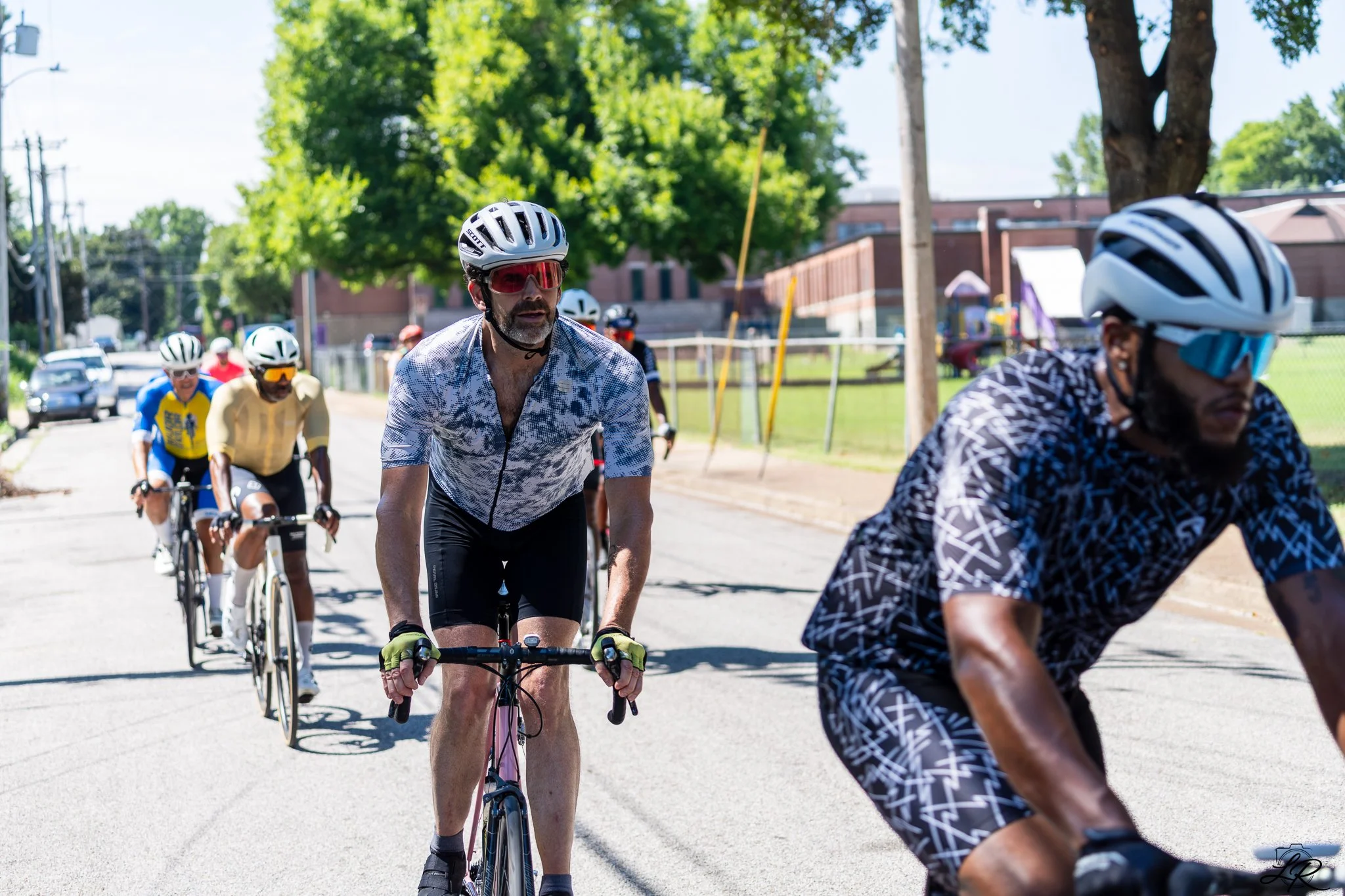 Group of people riding bicycles on a street, wearing helmets and sunglasses, with trees and buildings in the background on a sunny day.