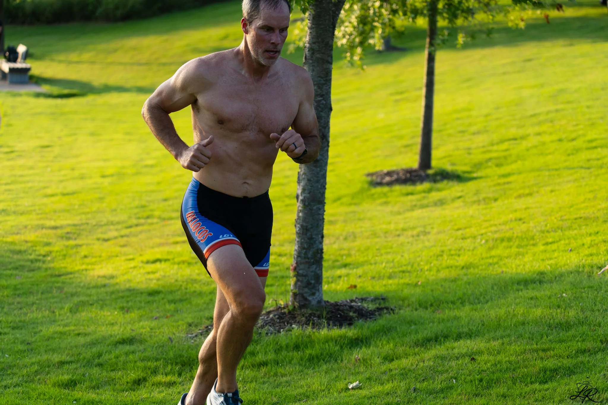 A shirtless man running outdoors on a grassy field with trees and a bench in the background.