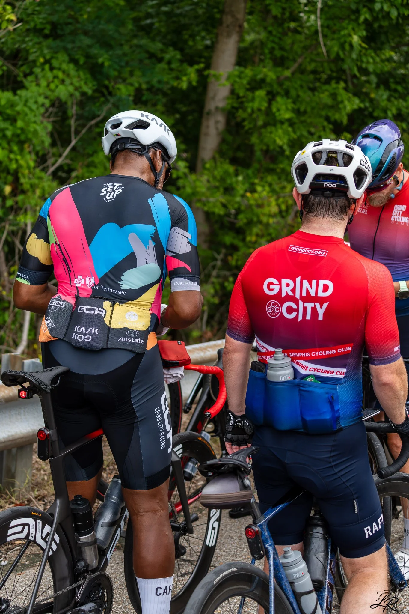 Three male cyclists in helmets and cycling gear gather outdoors near wooded area, with two bicycles visible.