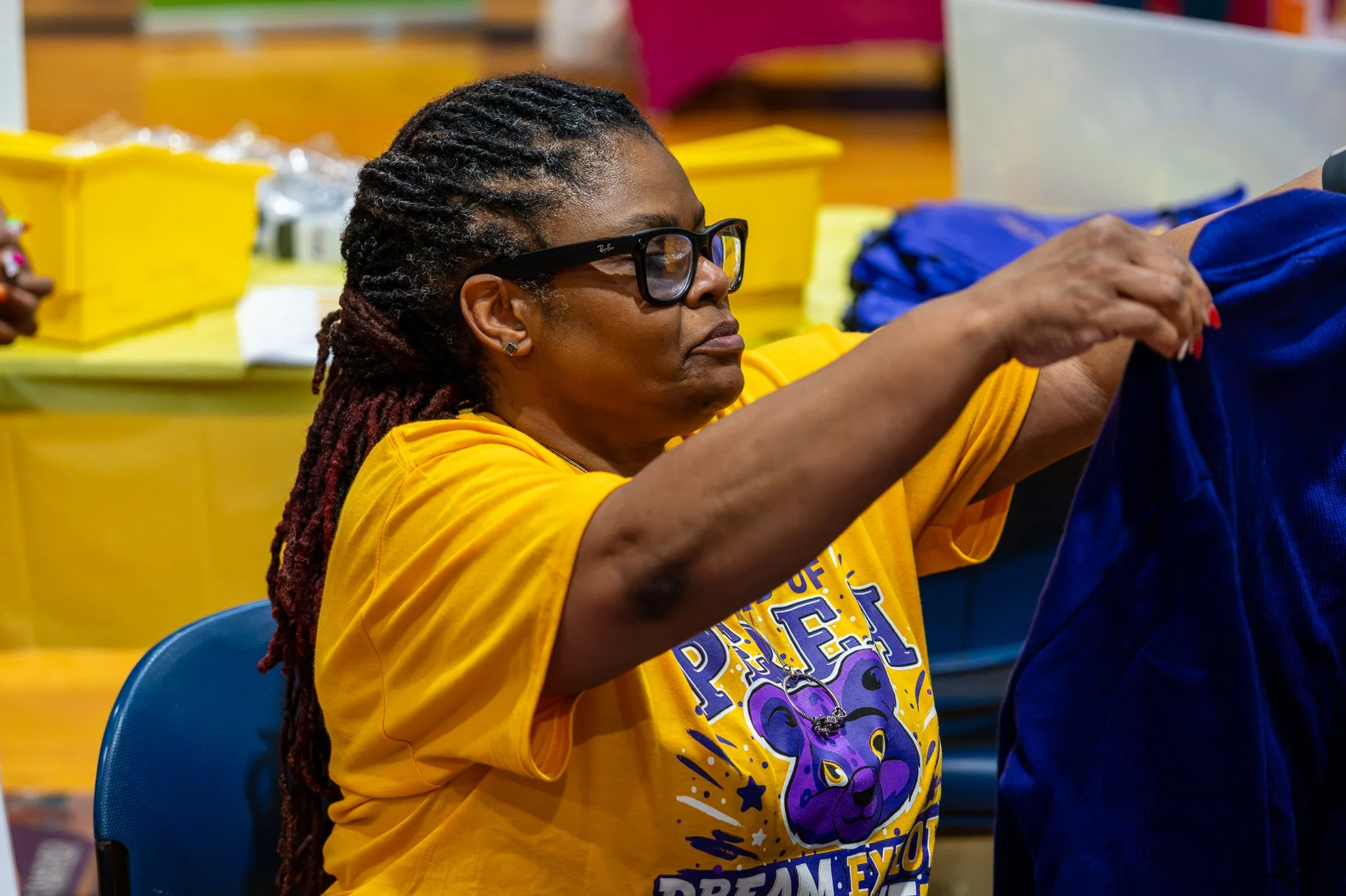 Woman with glasses and dreadlocks wearing a yellow T-shirt, sitting at a table in a room filled with yellow boxes and supplies.