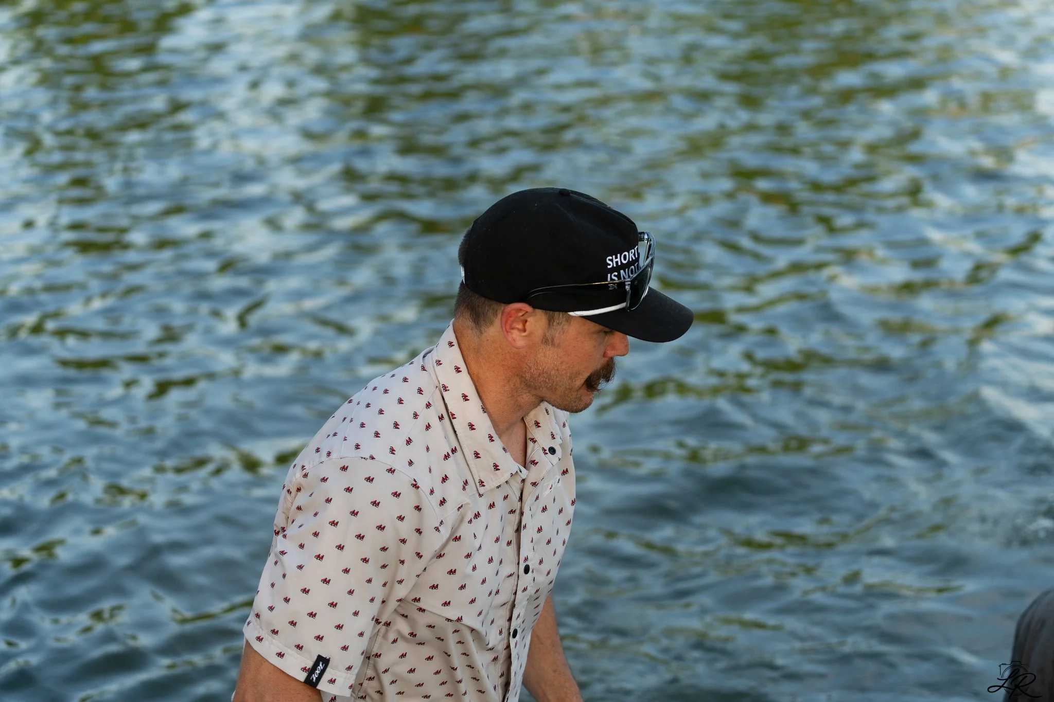 A man with light skin, short hair, and a mustache is wearing a black cap with white lettering and a patterned short-sleeve shirt. He is looking downward near a body of water with ripples.