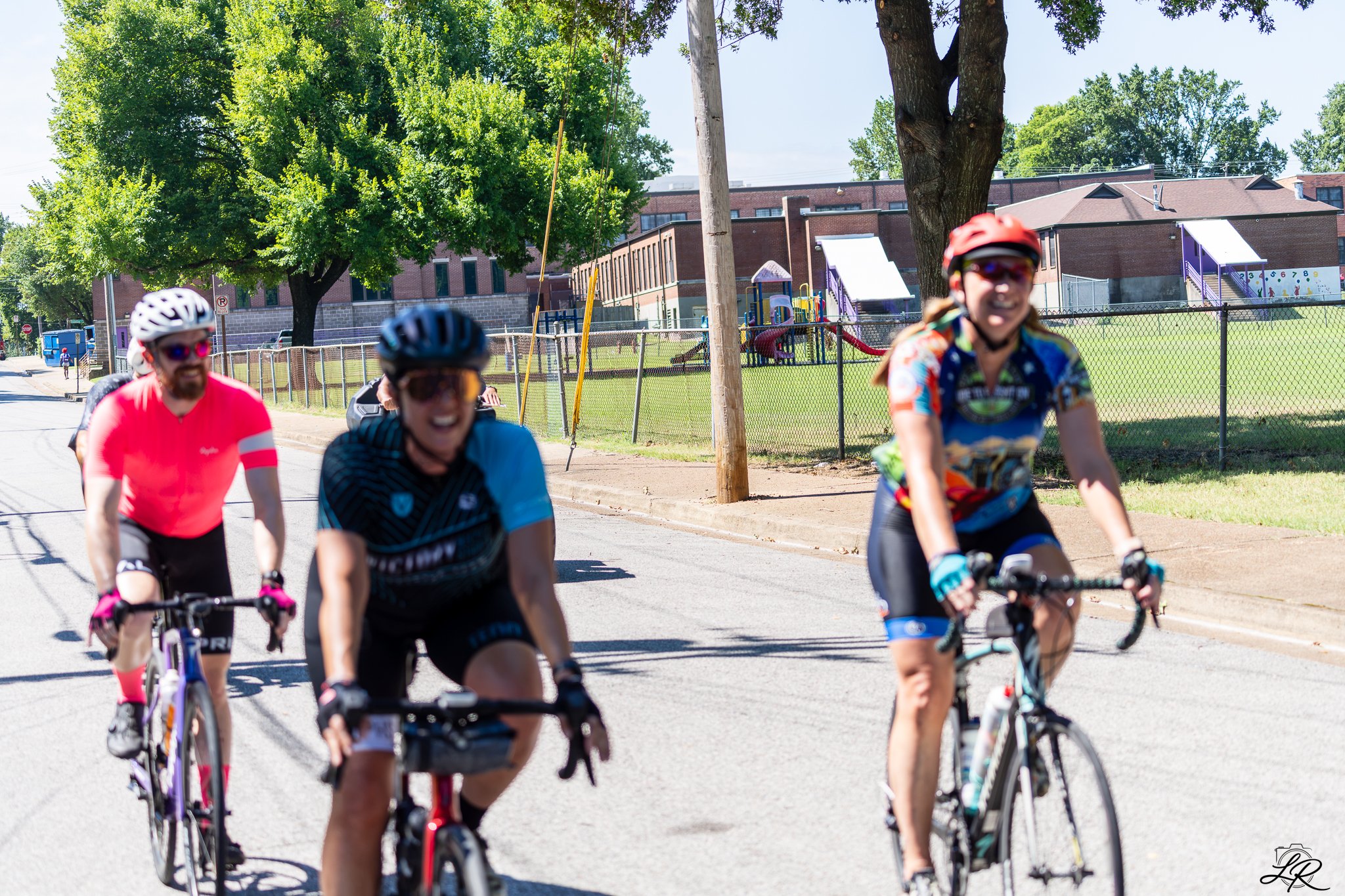 Group of four women riding bicycles on a sunny street, with a school building, playground, and large trees in the background.