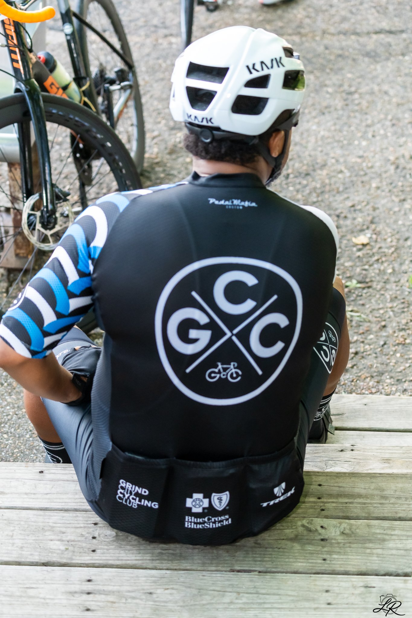 A person wearing a white bicycle helmet and black cycling jersey sitting on a wooden bench, with a bicycle and gravel ground in the background.