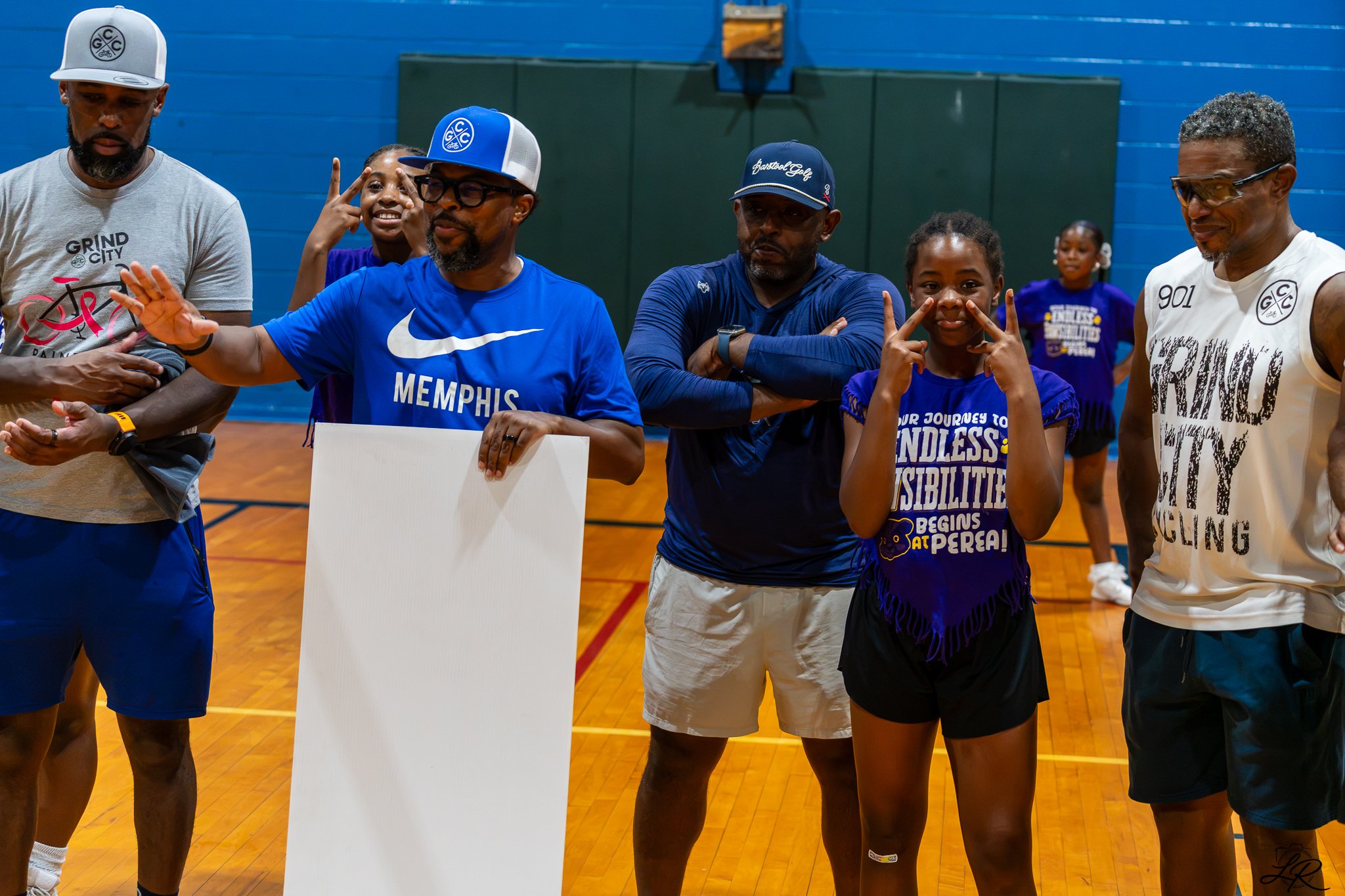 A group of men and girls standing on a basketball court indoors, some making peace signs and others listening.
