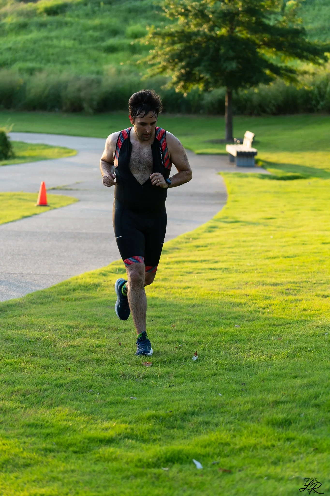 Man running outdoors on a paved path in a park, surrounded by green grass and trees, in the late afternoon.