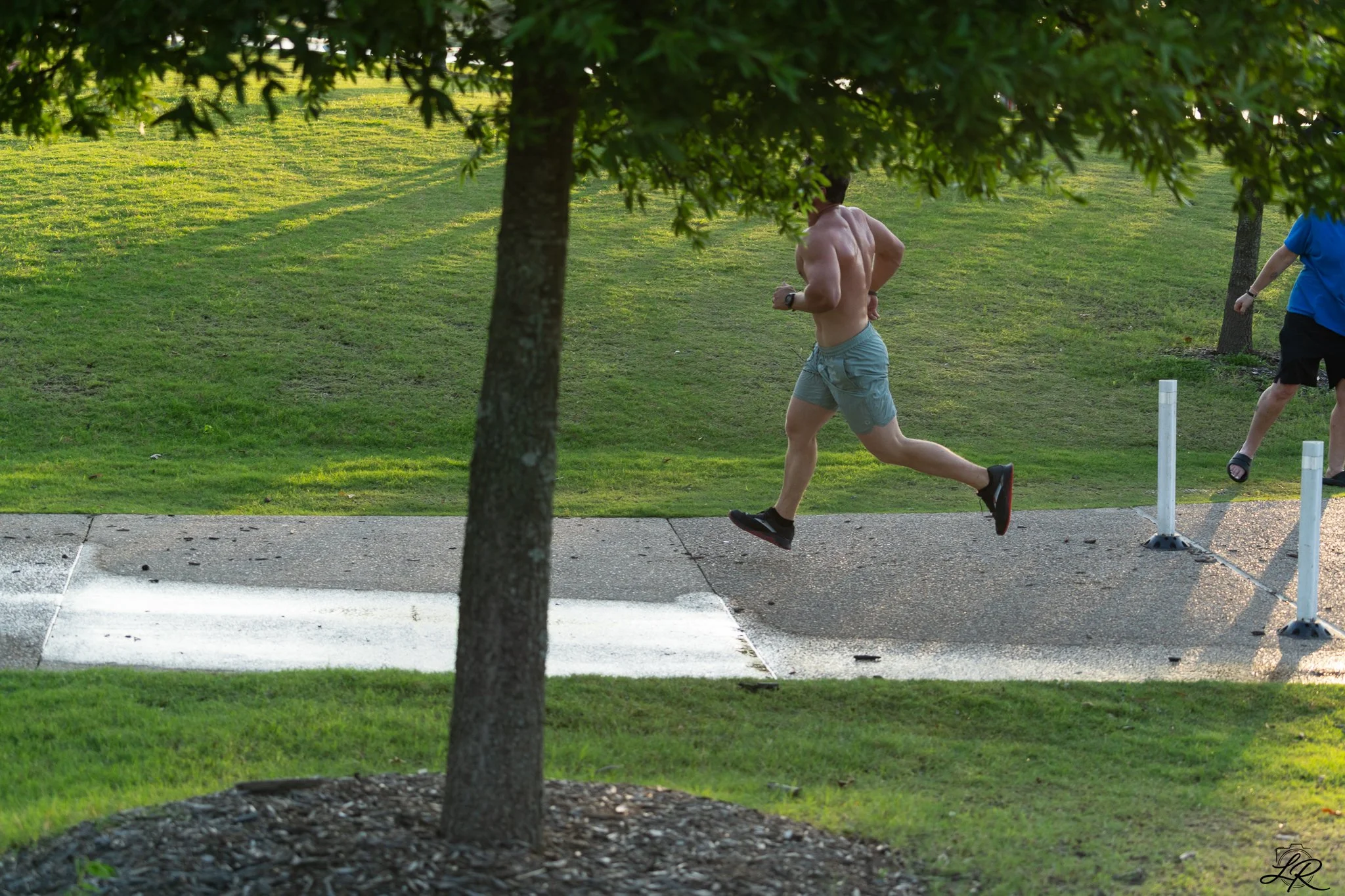 A shirtless man running outdoors on a grassy area, viewed from behind through a tree branch, with another person walking nearby, and trees and grass in the background.