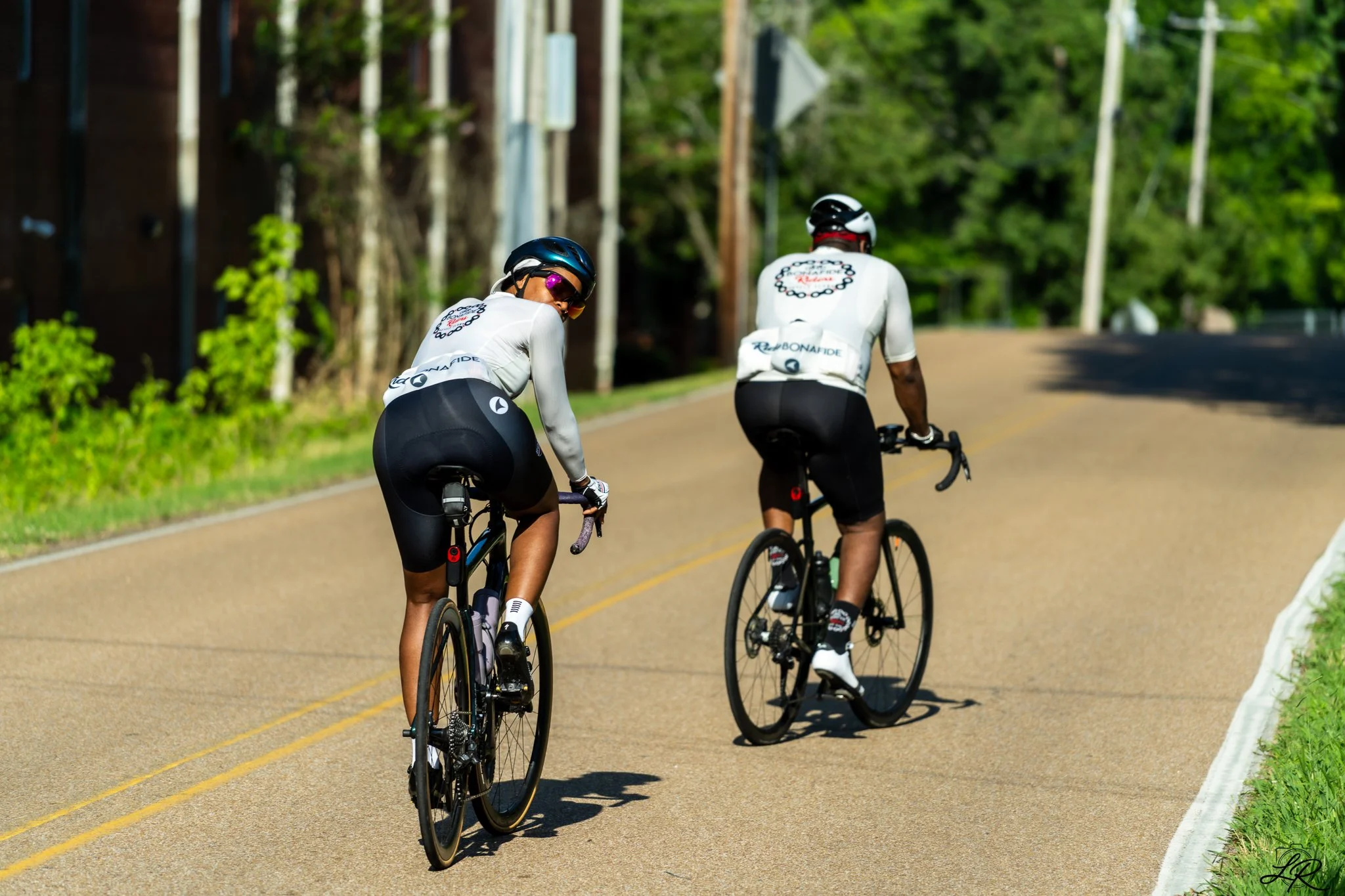 Two cyclists riding on a paved road surrounded by greenery and trees, with power poles and construction equipment visible in the background.
