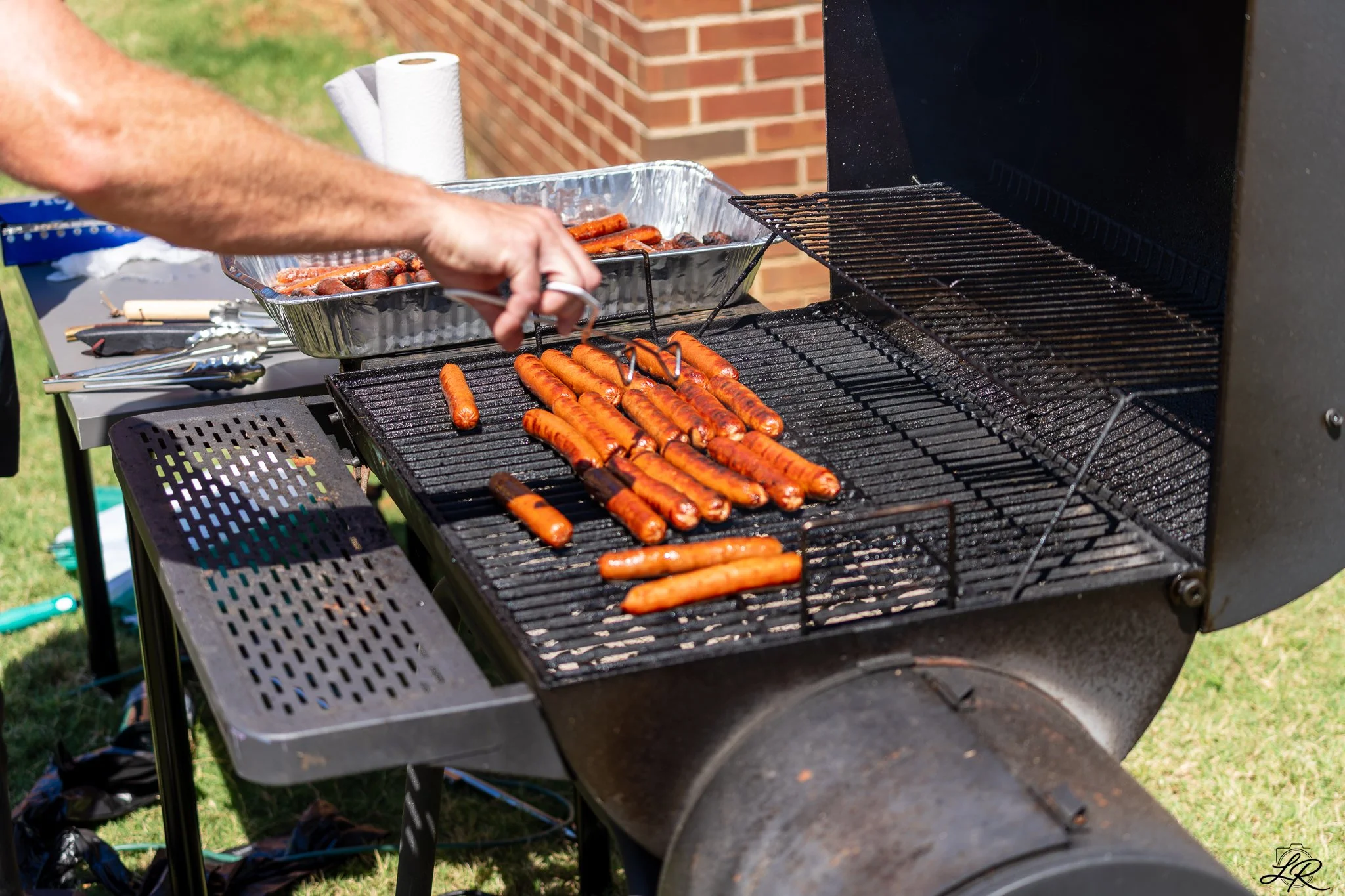 Sausages grilling on a barbecue grill outdoors with a brick wall in the background.