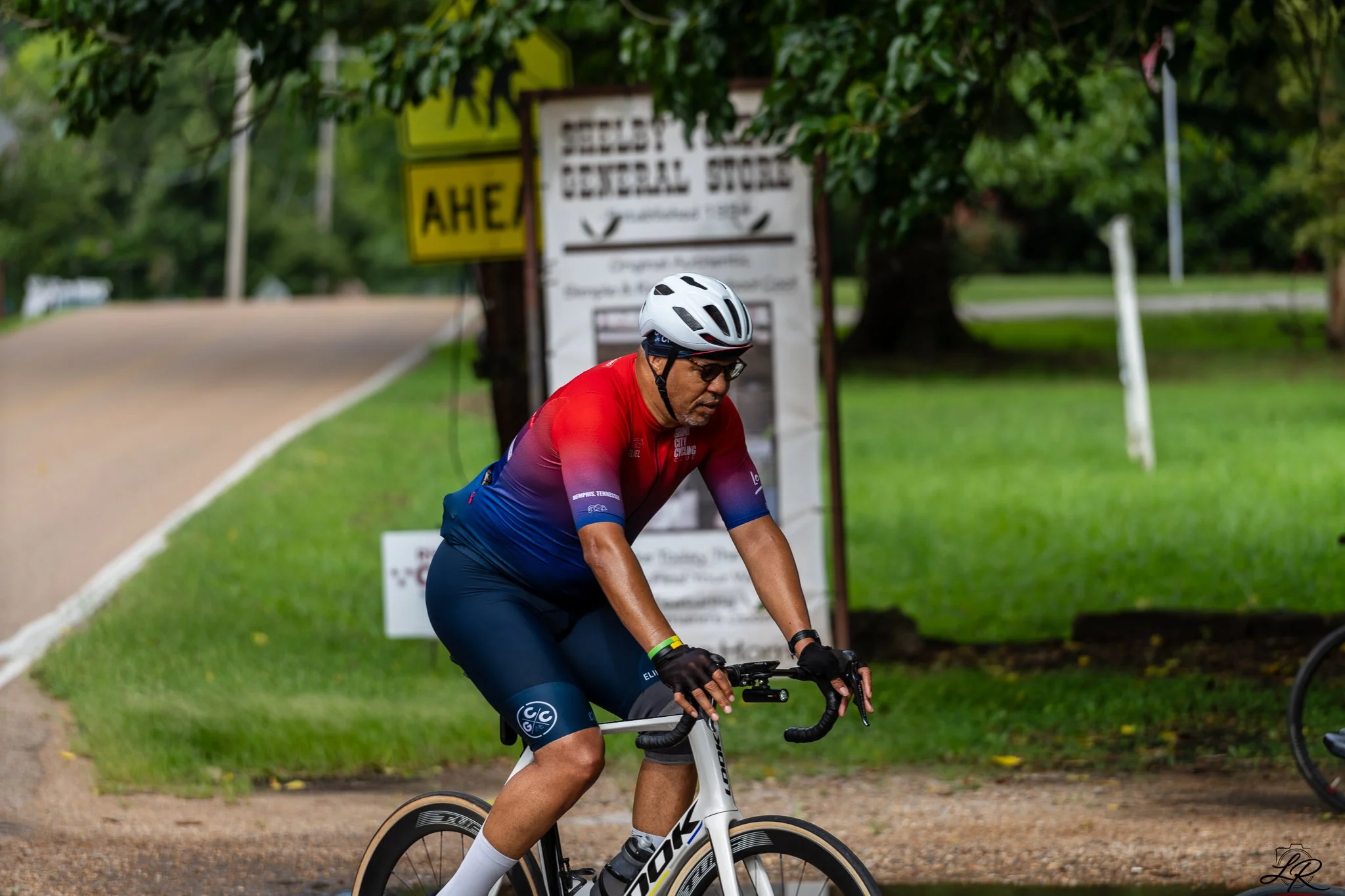 A man wearing a white helmet and red and blue cycling outfit riding a bicycle on a park path with green trees and a yellow sign in the background.