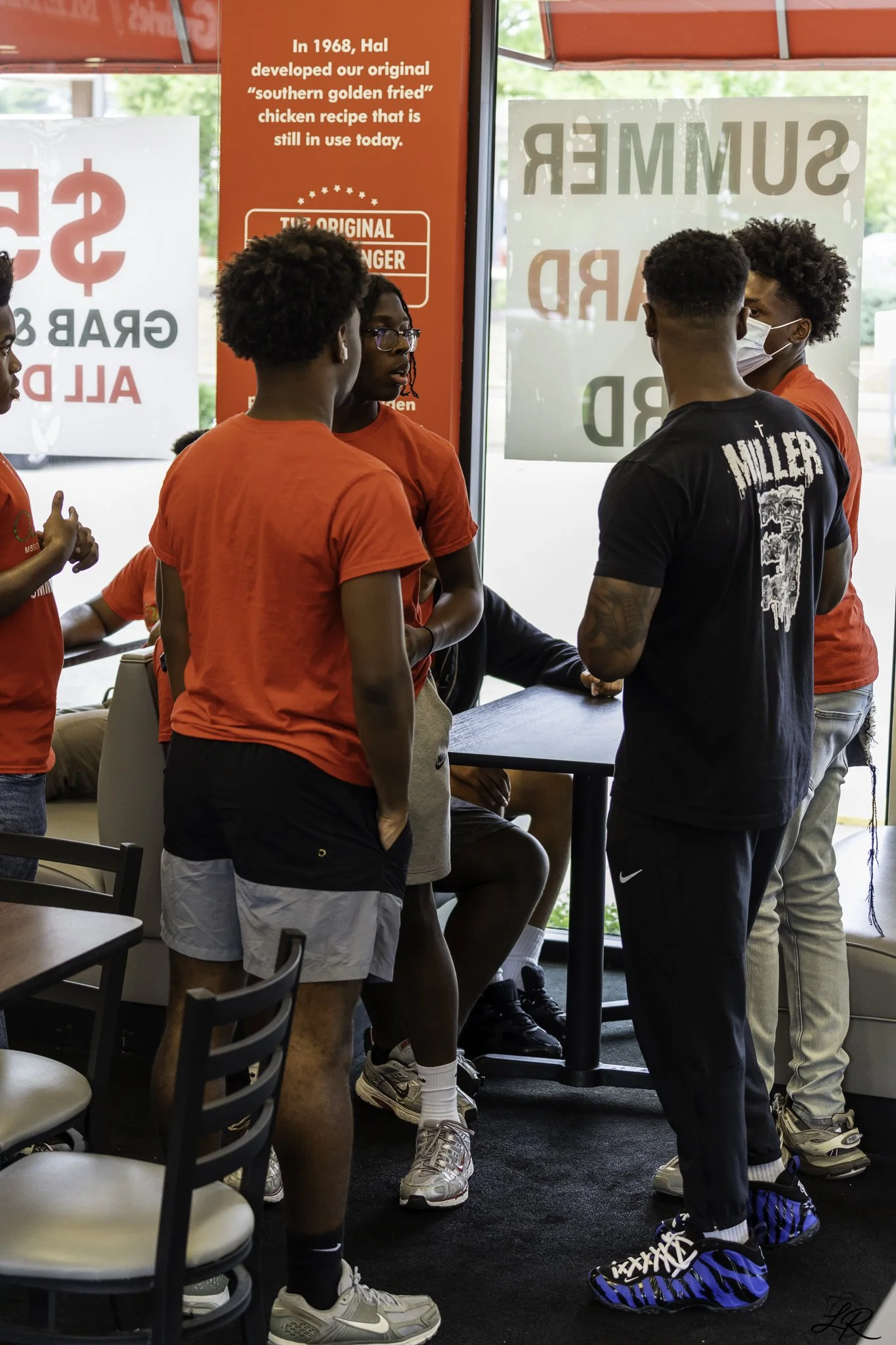 Four young men and one young woman are standing inside a restaurant, engaged in conversation. They are wearing athletic clothing and sneakers. Two of the men are wearing face masks. The background features red and white posters with text about a chic