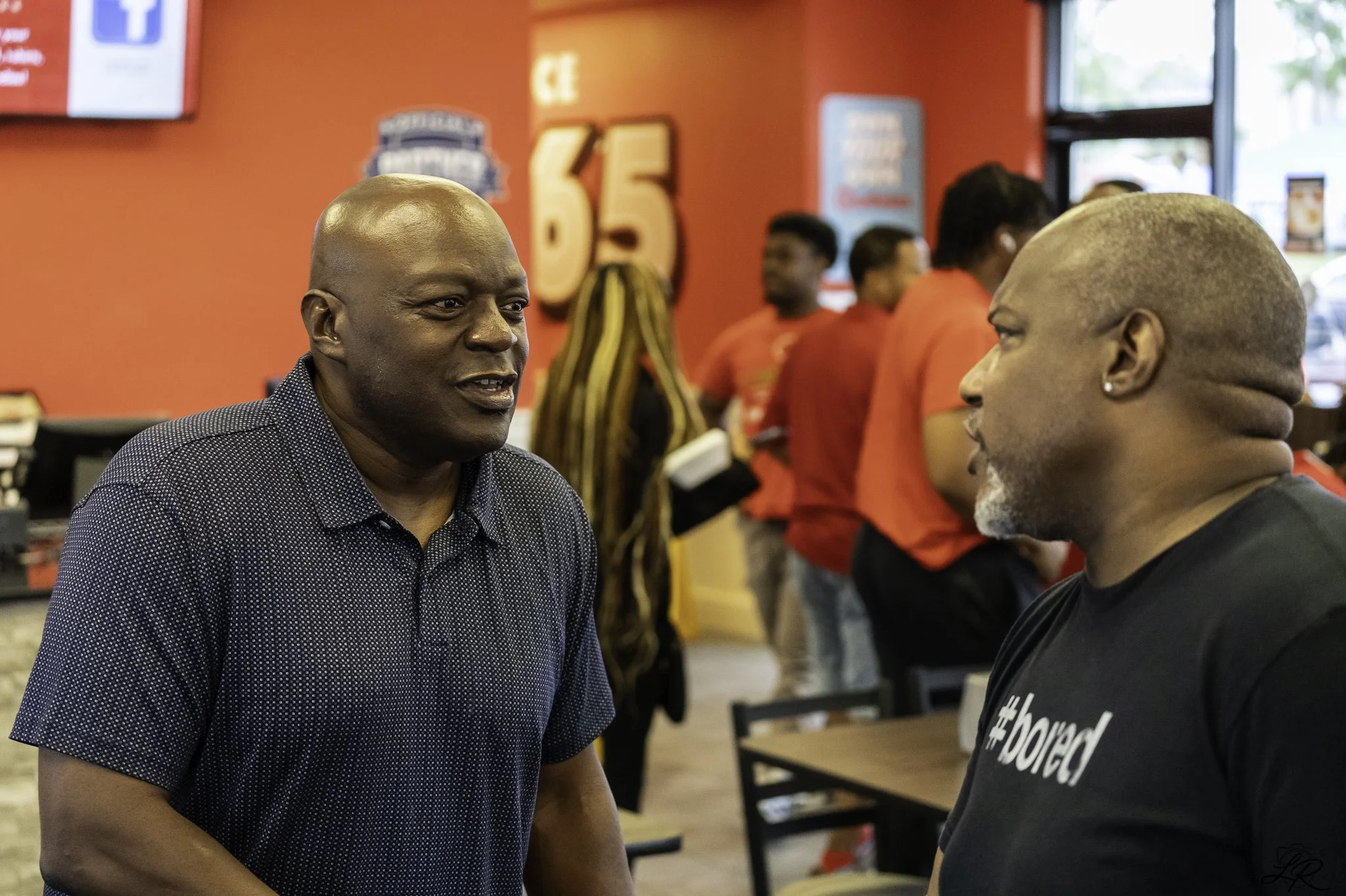 Two men engaged in conversation inside a restaurant with a red wall and people wearing red shirts in the background.