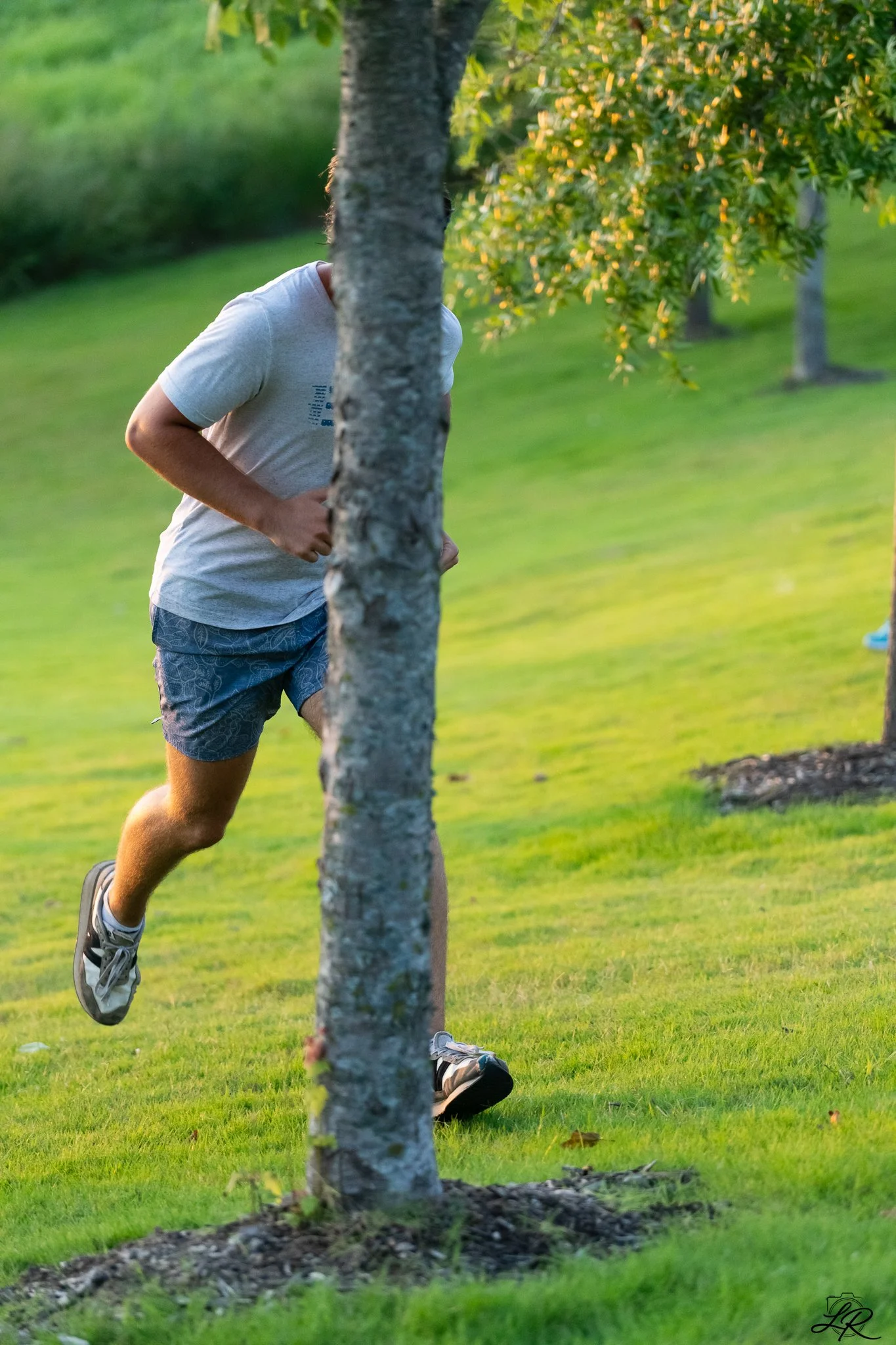 A person running outdoors on the grass, wearing a gray t-shirt, patterned shorts, and running shoes, partially obscured by a tree.