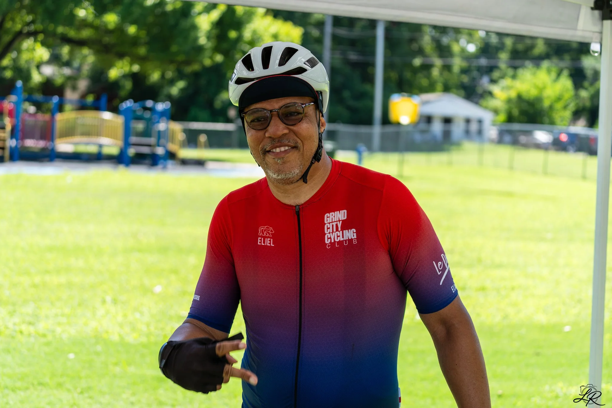 A man wearing glasses, a bicycle helmet, and a red cycling jersey with logos stands outdoors on a sunny day, smiling. There is green grass, trees, and some playground equipment in the background.