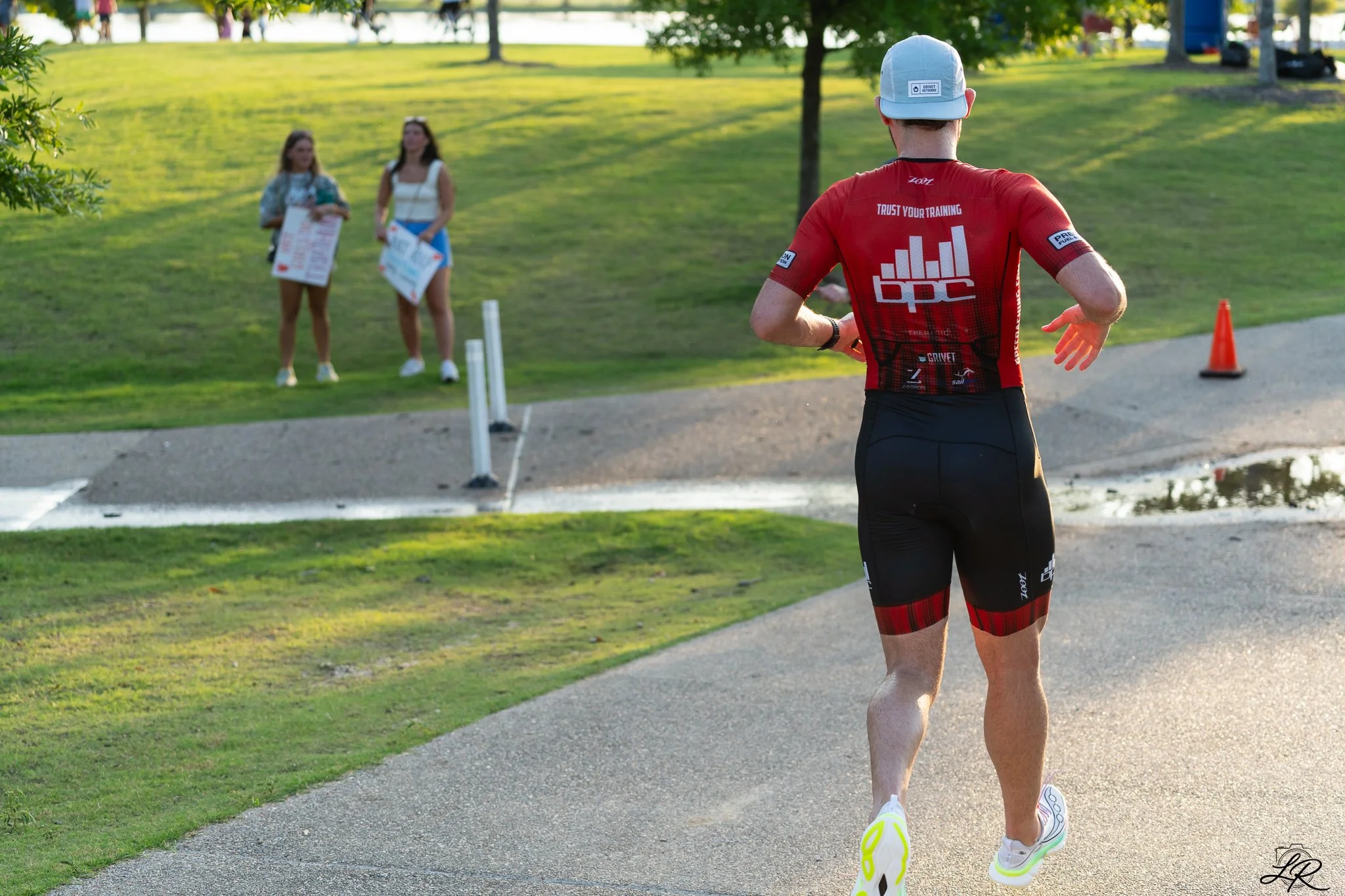 A man running outdoors in a park during sunset, wearing a red athletic shirt, black shorts, a gray hat, and bright yellow running shoes. Two women holding signs stand in the background on a grassy area surrounded by trees.