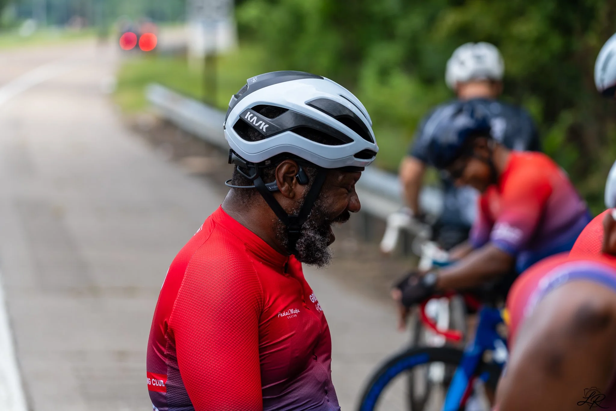 A man with a gray beard in a red cycling jersey and a black and white bike helmet, standing near a group of cyclists during a bike event.