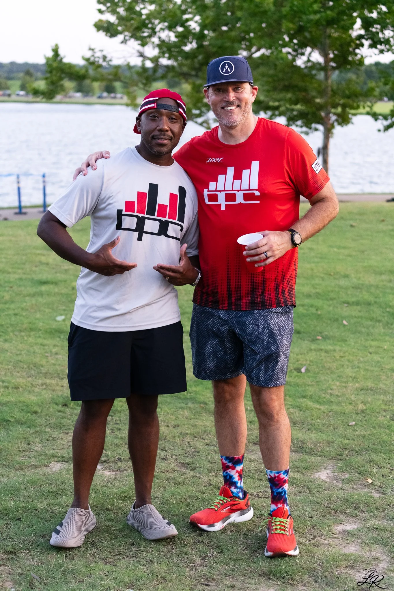 Two men standing outdoors near a lake or river, smiling and posing for the photo. One man wears a white T-shirt with a logo, black shorts, and gray shoes, while the other wears a red T-shirt with a logo, patterned shorts, colorful socks, and bright r