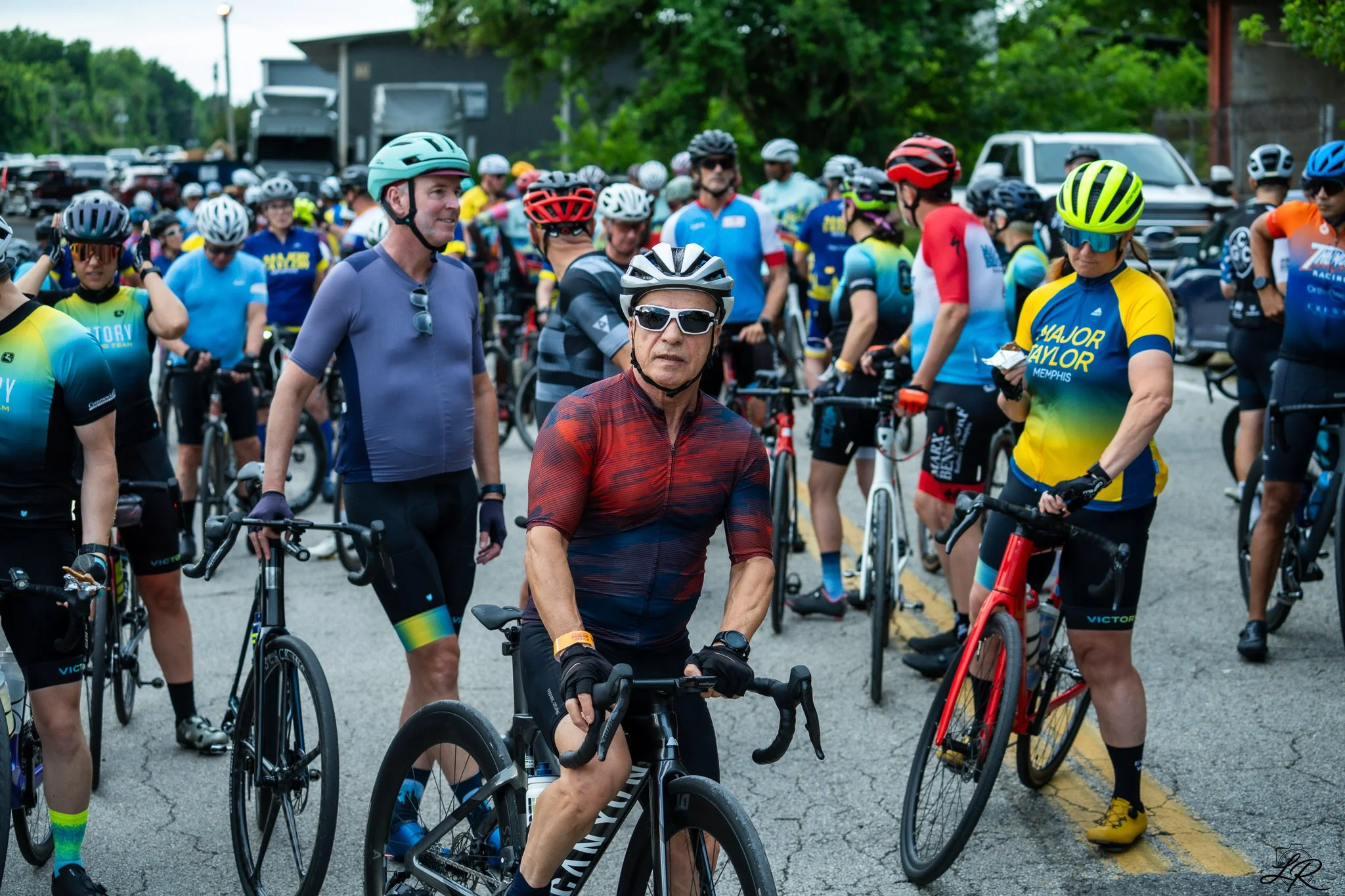 A group of cyclists gathered outdoors, preparing for or taking part in a cycling event. They are wearing helmets and sporting colorful cycling jerseys and gear.