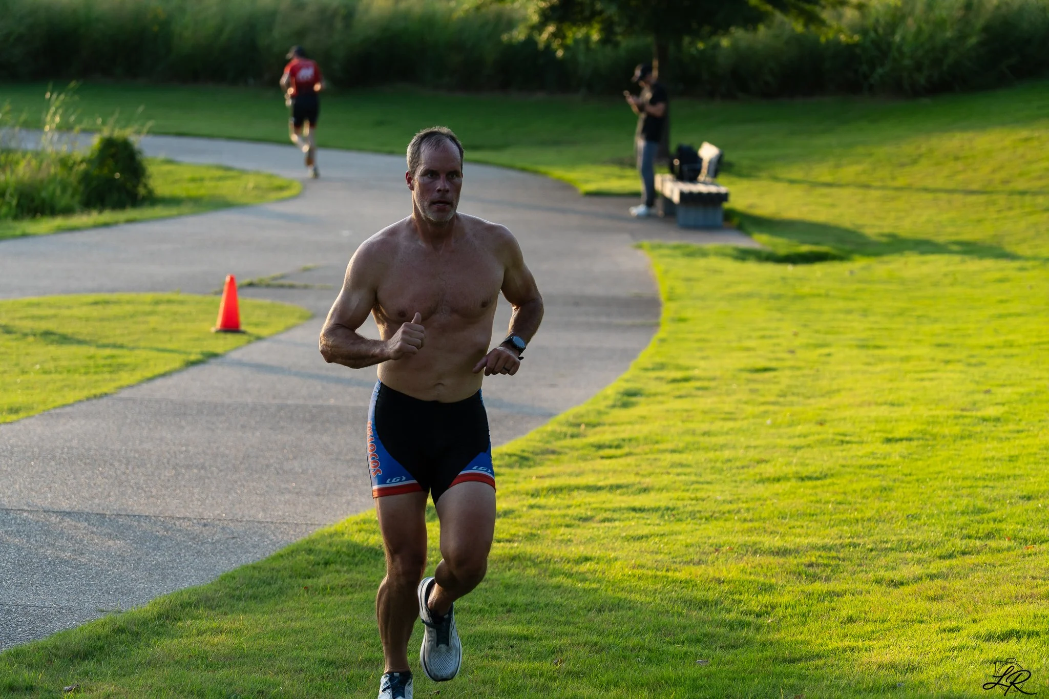 A shirtless man running outdoors on a paved path in a park area with green grass, trees, and two other people in the background. One person is jogging, and the other is standing and using a phone.