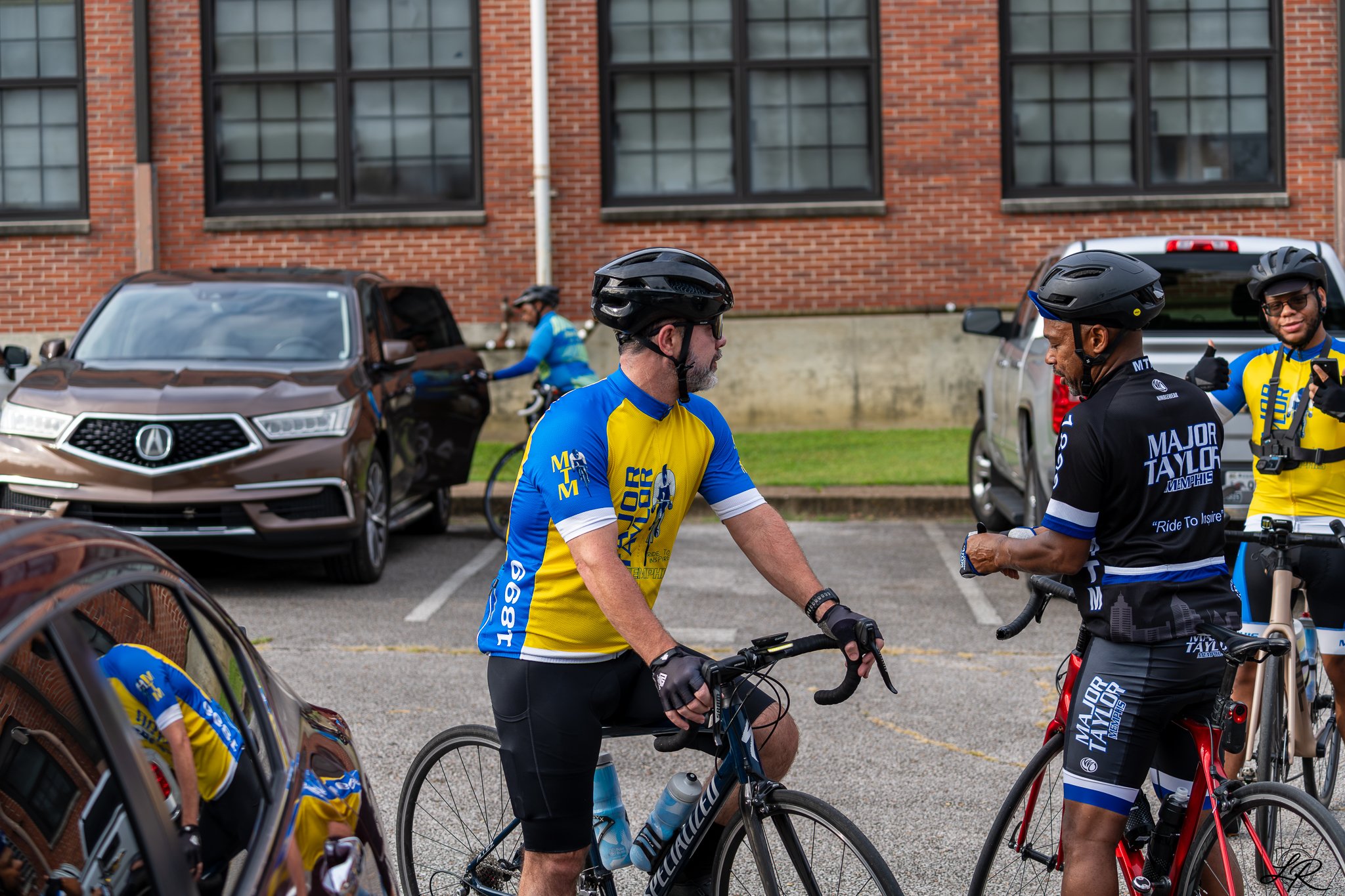 Cyclists in colorful jerseys and helmets gathered in a parking lot, with brick building and cars in the background.
