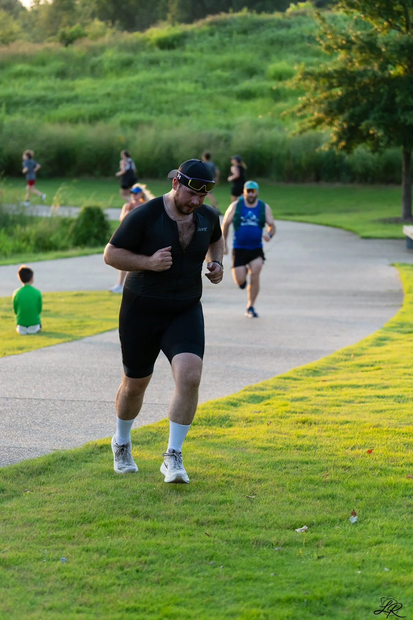 A man in athletic clothing running on a paved trail through a park with green grass, trees, and a hillside in the background, during late afternoon or early evening.