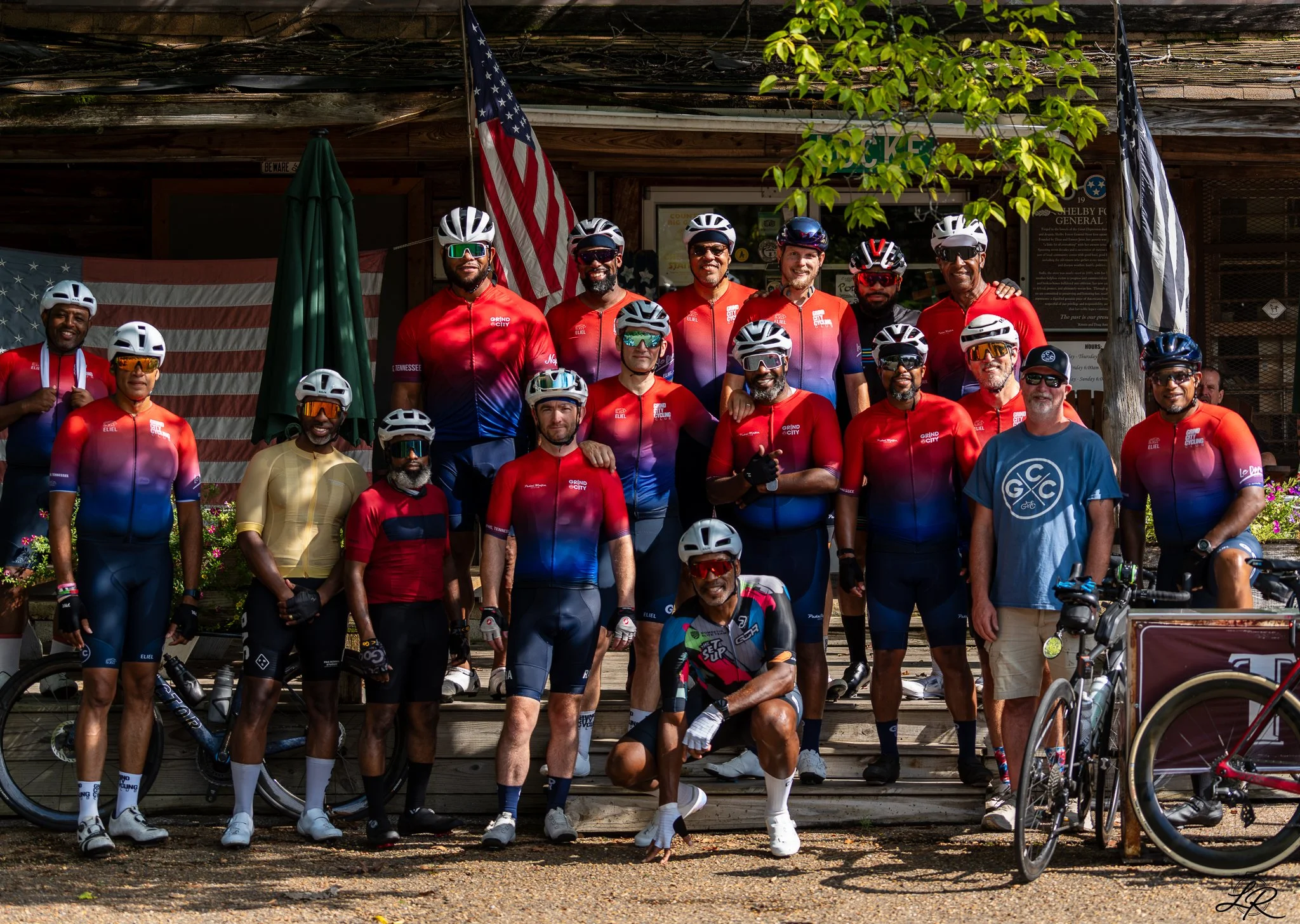 A group of cyclists wearing matching red and purple jerseys, helmets, and sunglasses standing on wooden steps in front of a building with American flags. There are palm trees and bicycles around them.