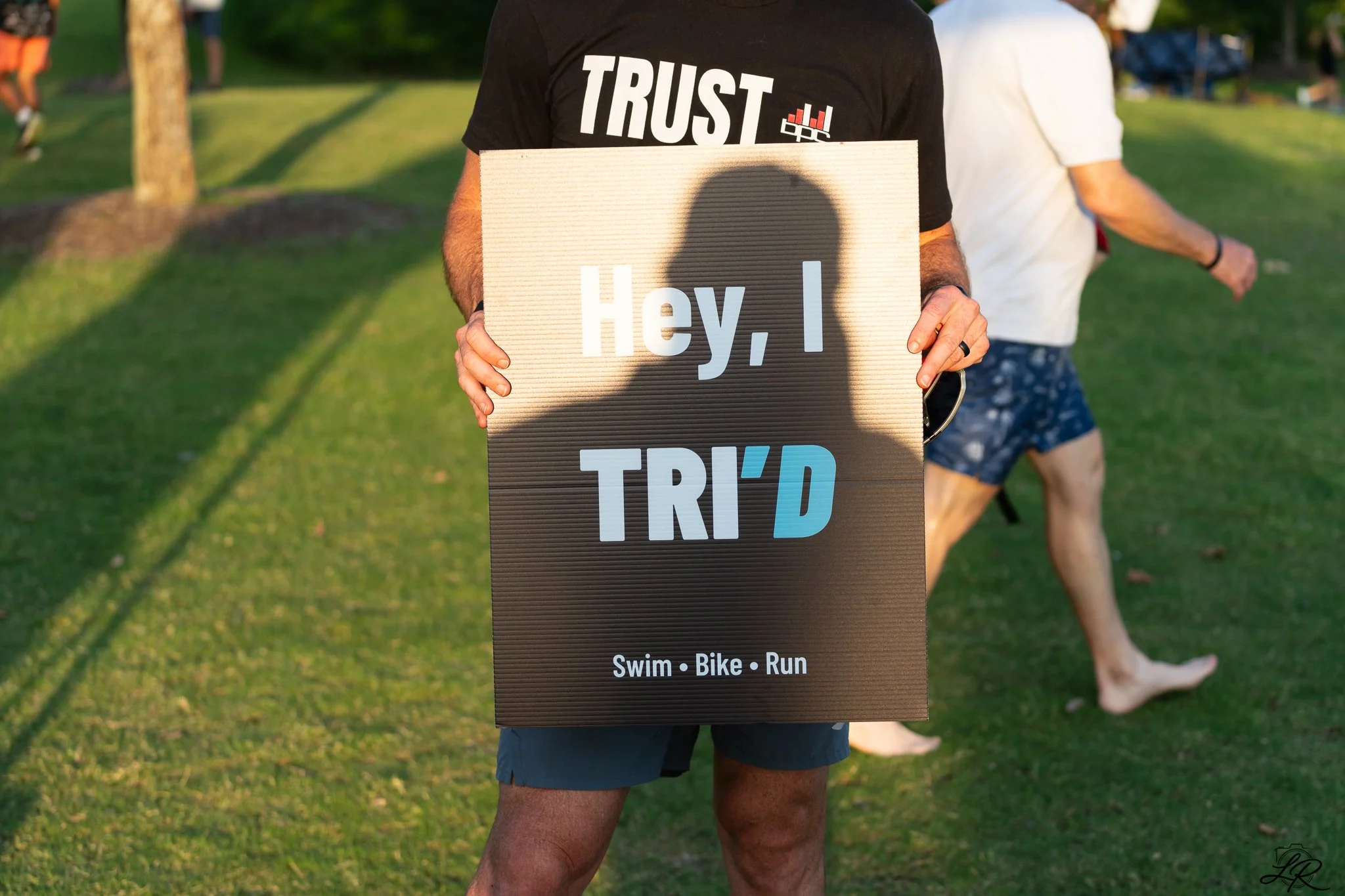 Person holding a sign with the text "Hey, I' D" and the words "Swim, Bike, Run" underneath, outdoors on a grassy area with trees and other people in the background.