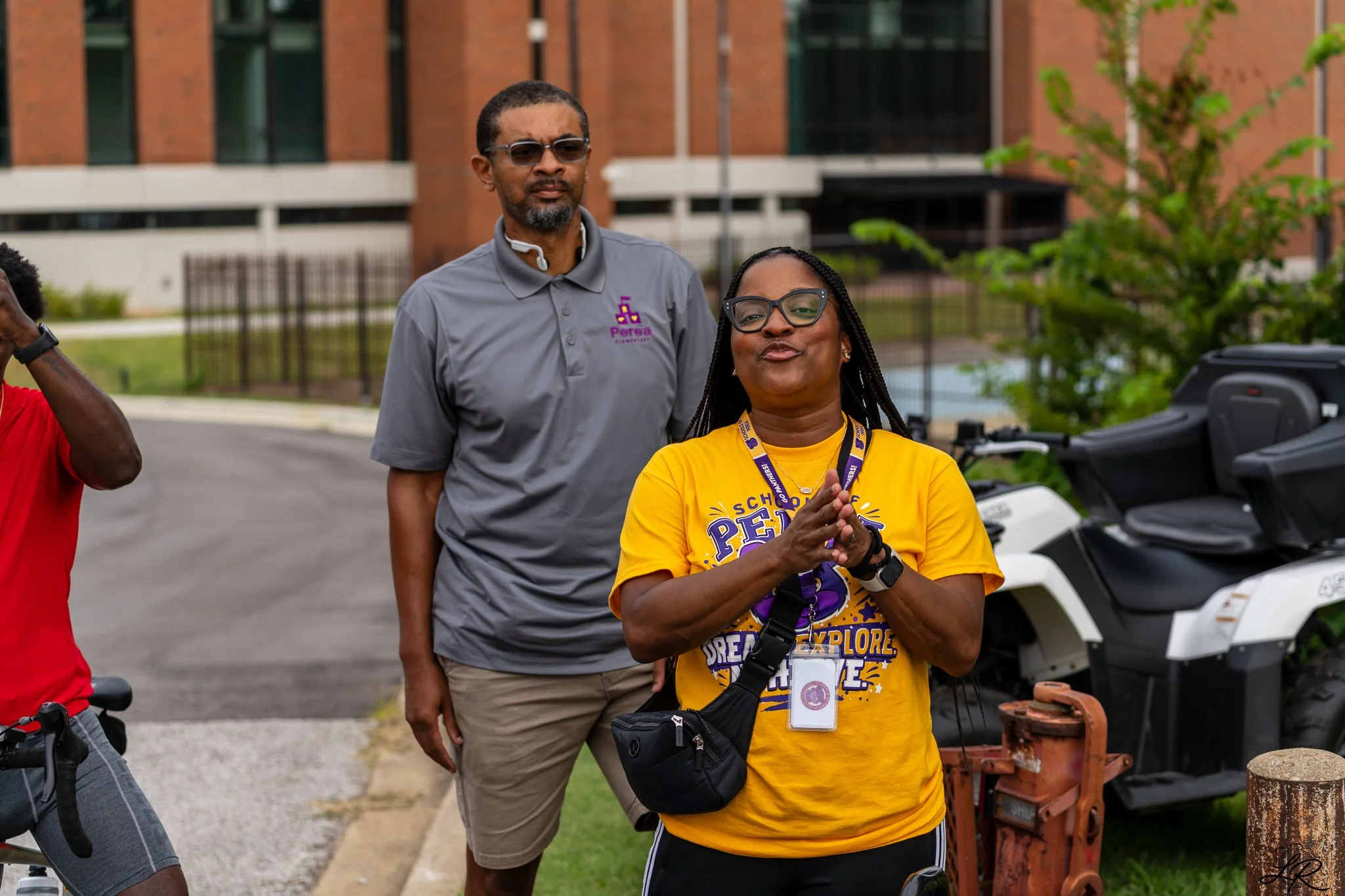 Group of people outdoors, woman in yellow shirt with glasses, woman in gray shirt with logo, man with sunglasses, biking gear, standing near a golf cart, trees and building in background.