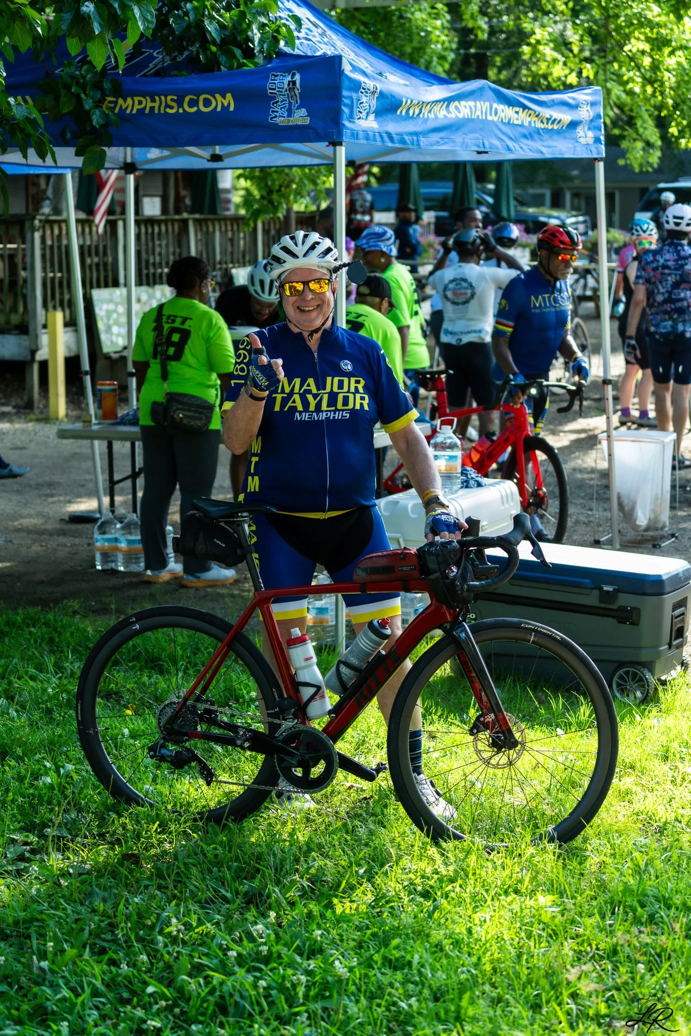 Smiling cyclist wearing glasses, a blue and yellow "Major Taylor Memphis" jersey, and a white helmet, standing with a red bicycle under a tree at a cycling event with multiple participants and a blue tent in the background.