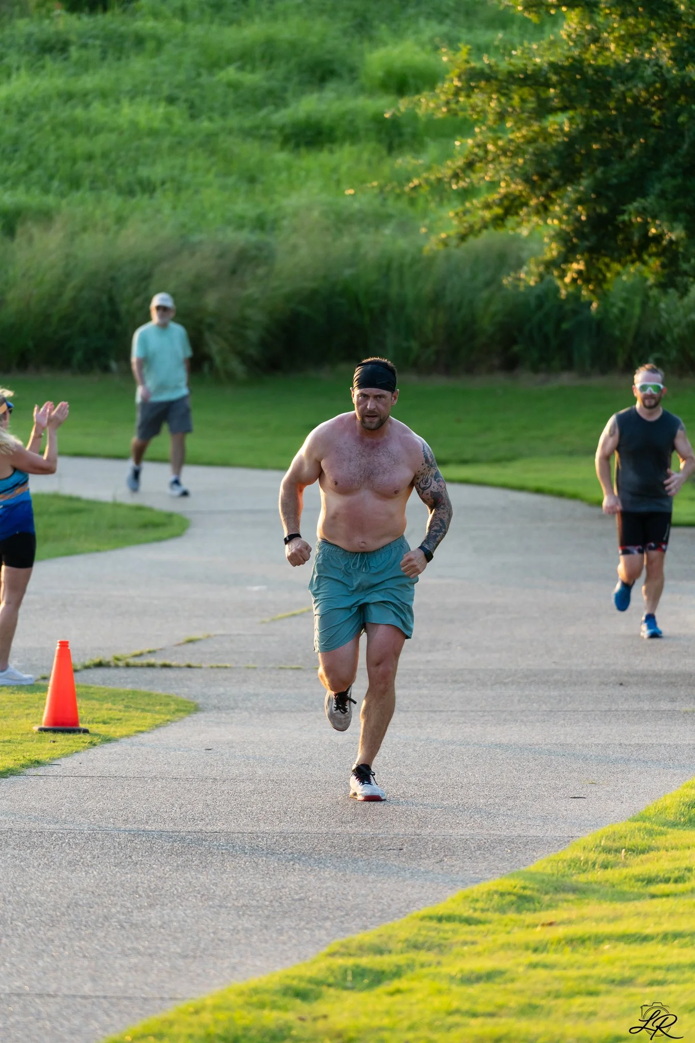 A shirtless man running on a paved path in a park, with an orange cone on the side, and several people observing or jogging nearby, green grass and trees in the background.