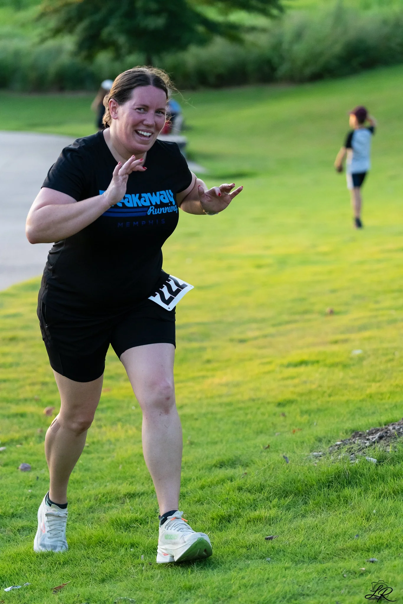 A woman running outdoors on a grassy field, smiling, wearing a black t-shirt and shorts, with other people in the background.