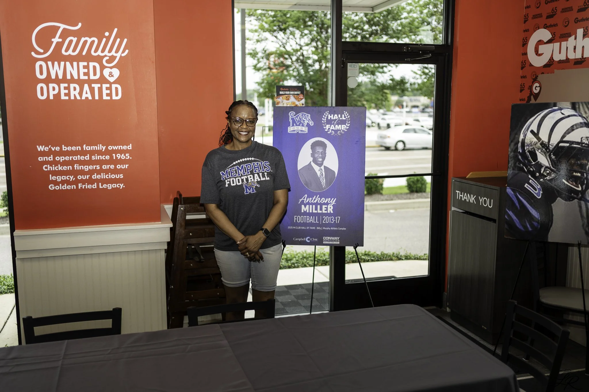 A woman standing indoors in front of a sign that reads 'Memphis Football Hall of Fame' with a portrait of a man named Anthony Miller. The woman is wearing a Memphis Football t-shirt, glasses, and smiling. There is a red wall with text about the famil