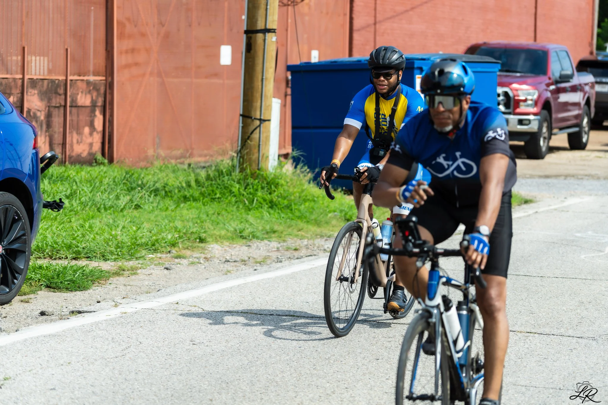 Two men riding bikes on a road, both wearing helmets, sunglasses, and cycling jerseys, with a red truck, blue container, and parked cars in the background.
