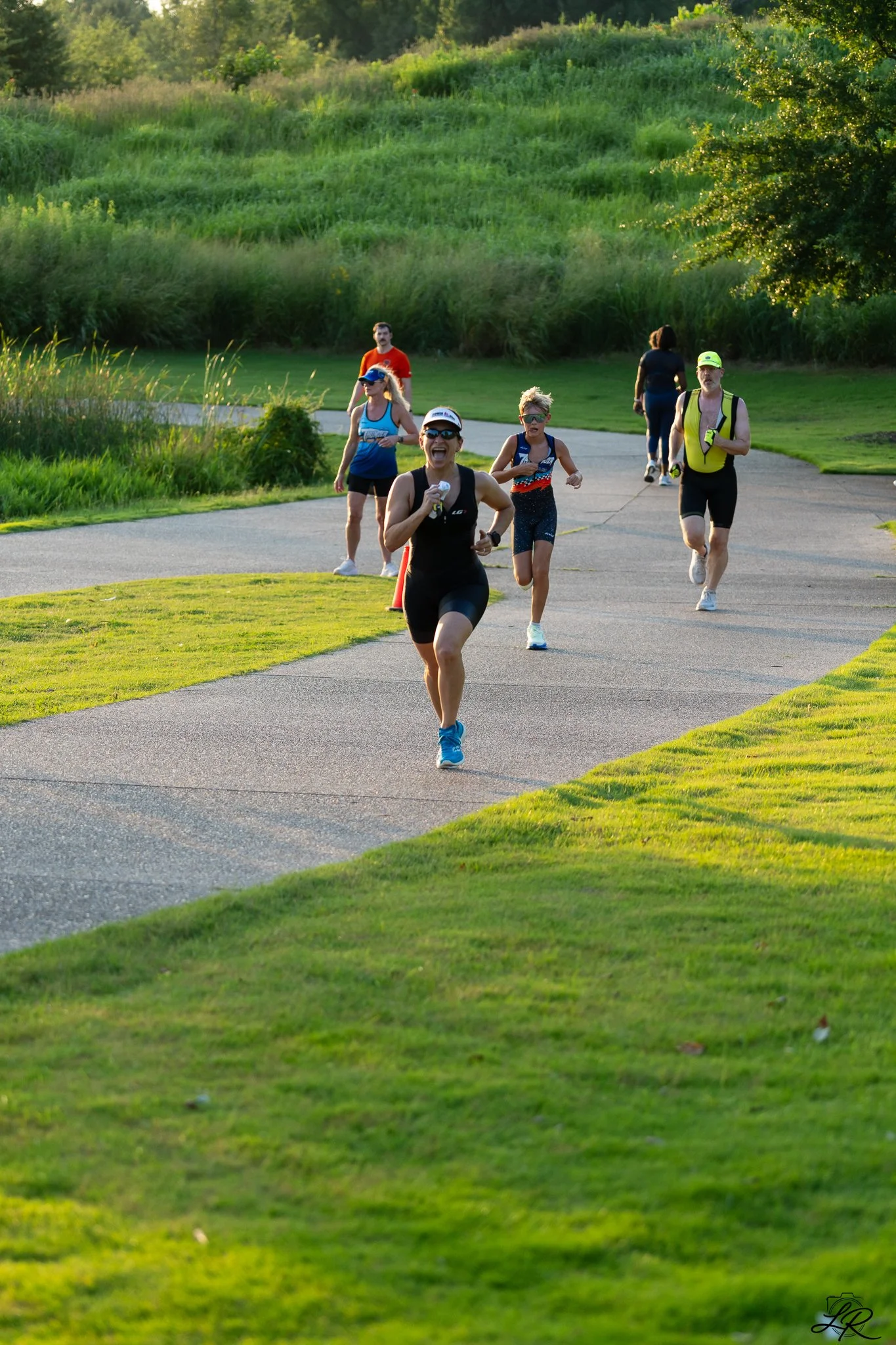 A group of six people running or jogging on a paved trail in a park with lush green grass, trees, and a grassy hill in the background during a sunny day.