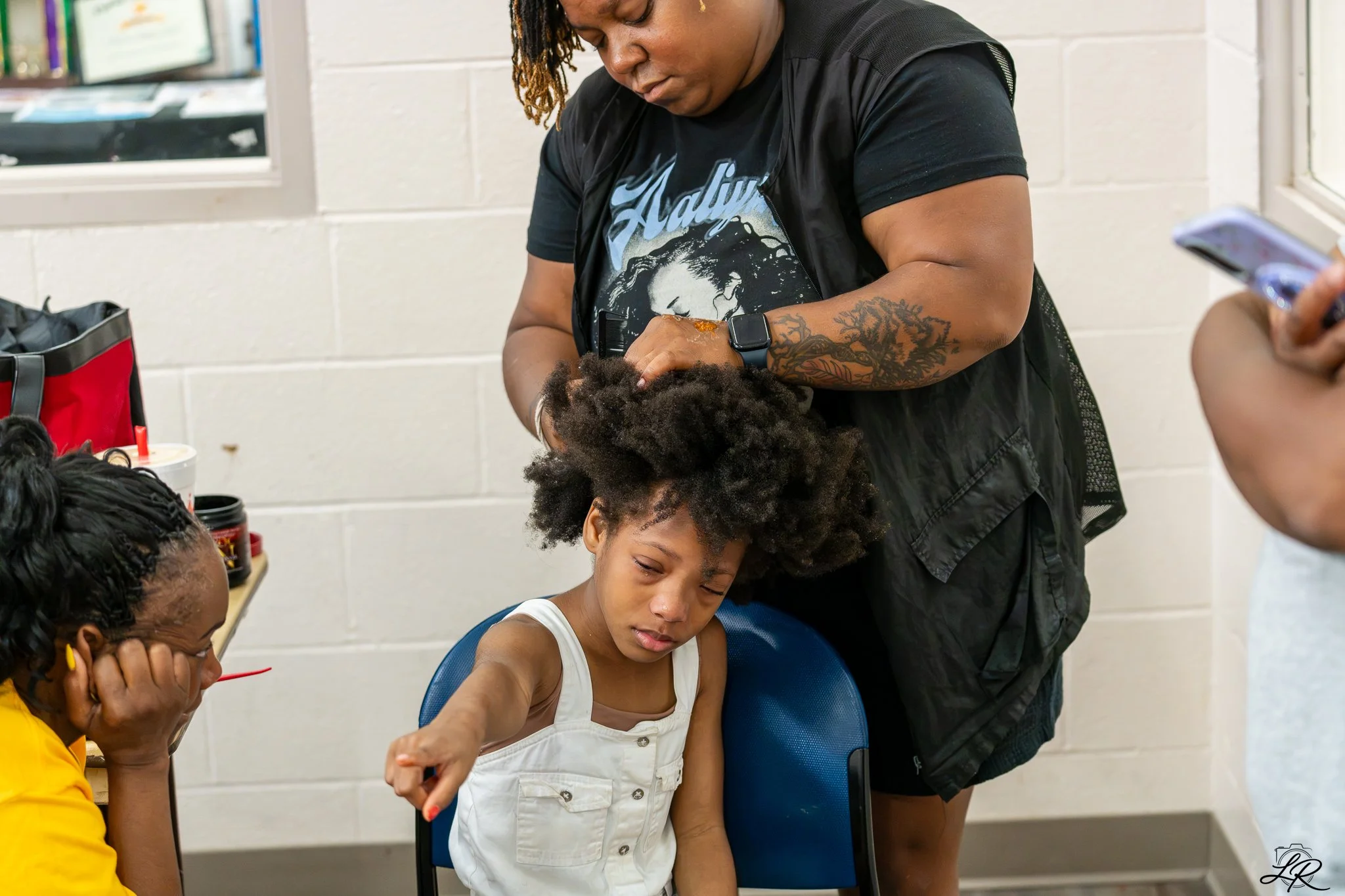 A woman with a tattoo on her arm is styling a young girl's hair, while other women watch in a classroom or community center with white brick walls and windows.