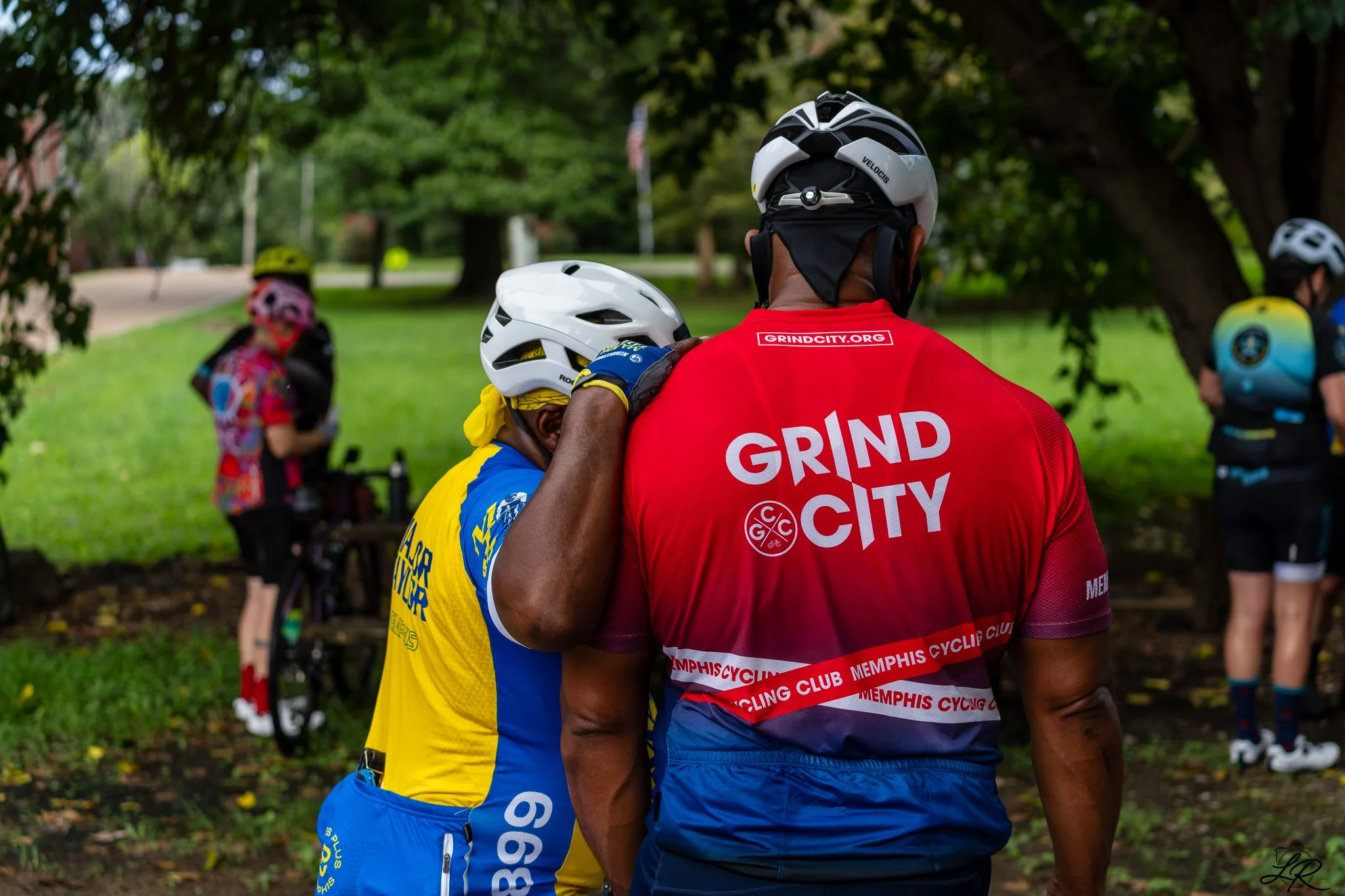 Two cyclists, one wearing a red jersey with 'GRIND CITY' and 'MEMPHIS CYCLING CLUB' written on it and the other in yellow and blue, are embracing each other under trees in a park, with other cyclists and bikes in the background.