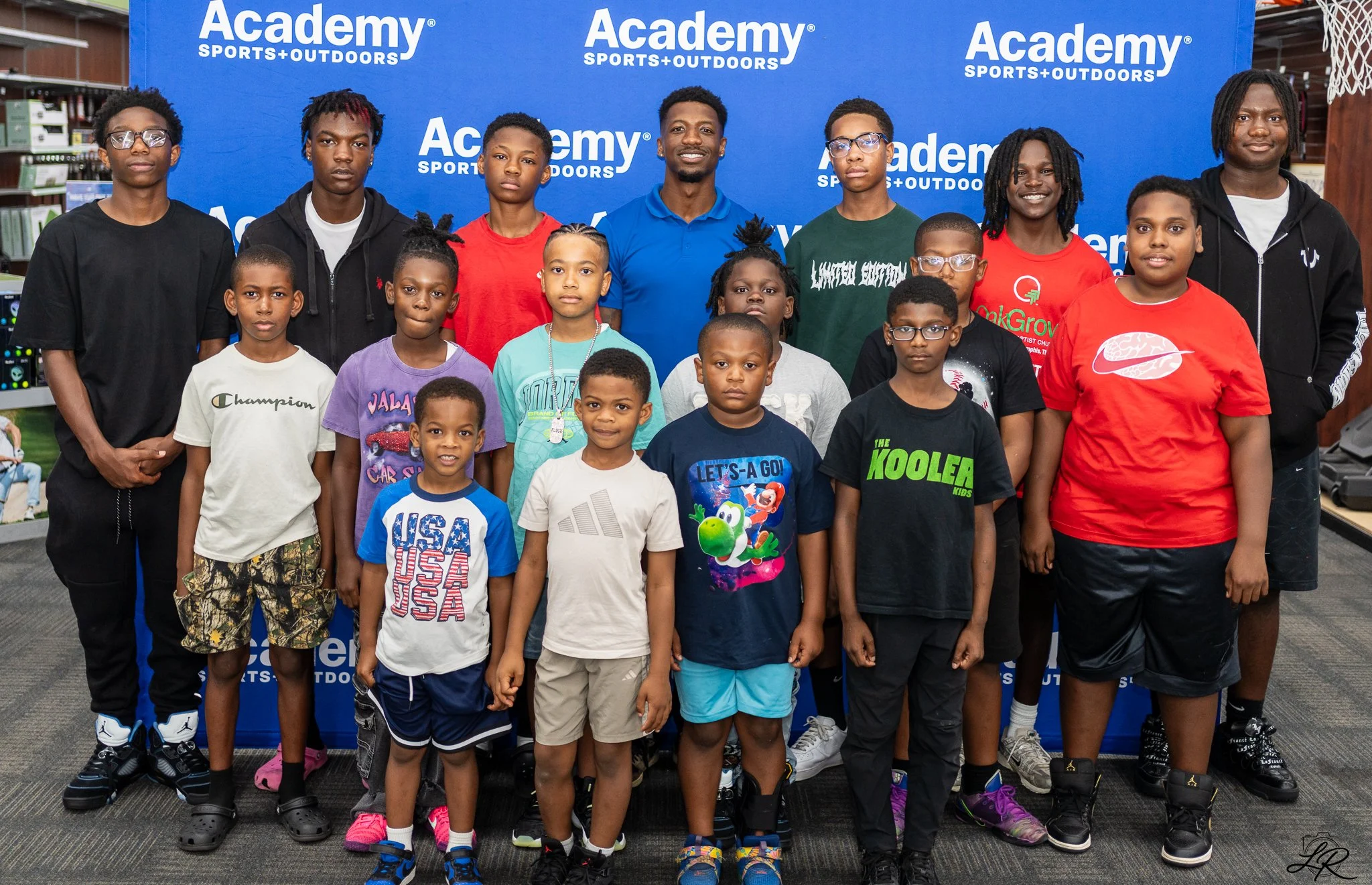 Group of children and teenagers posing in front of a blue backdrop with 'Academy Sports + Outdoors' logos, inside a store.