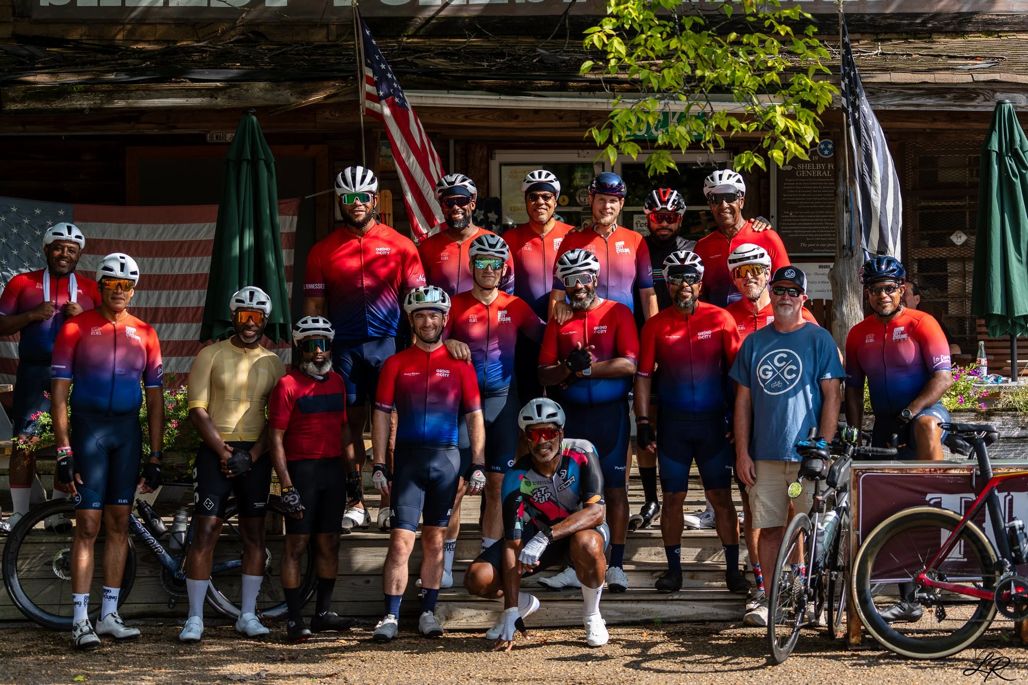 Group of cyclists wearing helmets and colorful cycling uniforms posing in front of a rustic building decorated with American flags.