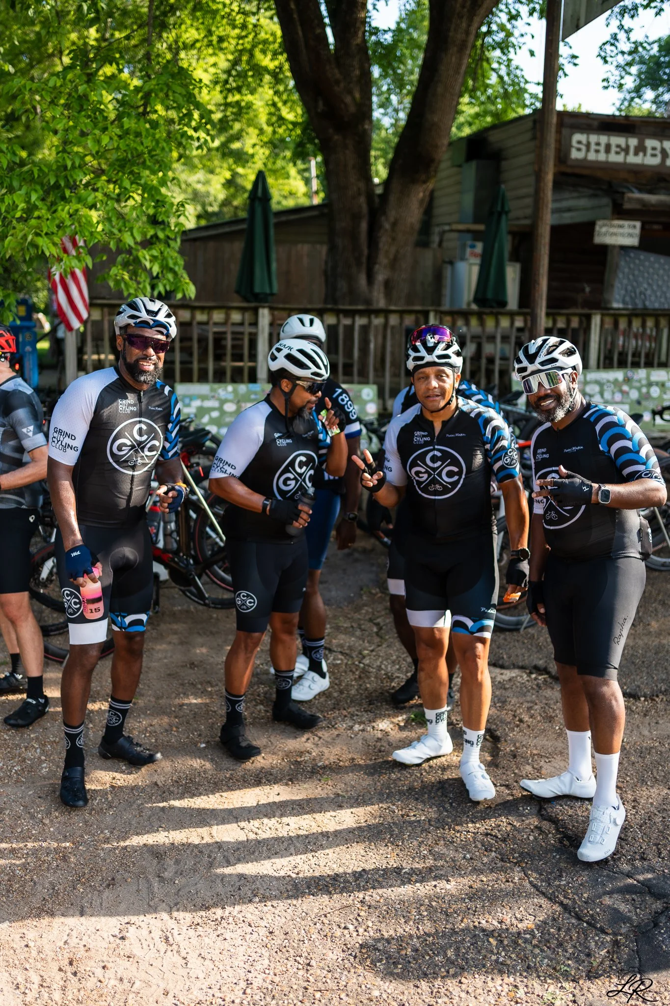A group of five cyclists wearing matching black, white, and blue cycling jerseys and helmets, standing outdoors on a dirt path beneath a large tree with green leaves, with a wooden fence, umbrellas, and a building with a sign in the background.