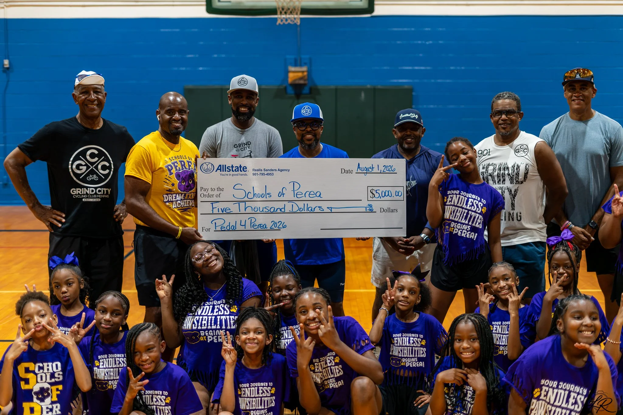 Group of children and adults in gymnasium, holding oversized check for $5,000 from Allstate for Schools of Perea, with children wearing purple shirts.