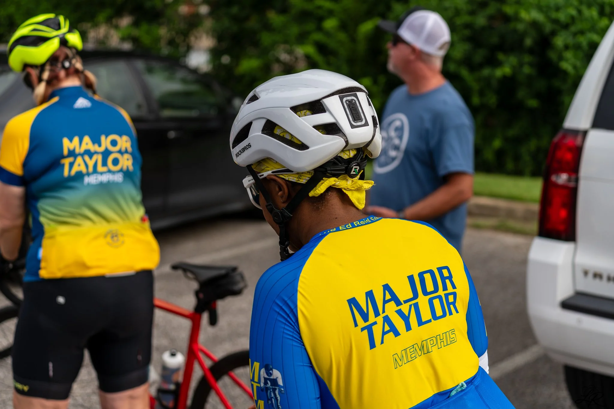 A group of cyclists wearing blue and yellow jerseys with 'Major Taylor Memphis' written on the back, gathered outdoors with a man in a blue shirt and white cap, and cycling helmets visible in the scene.