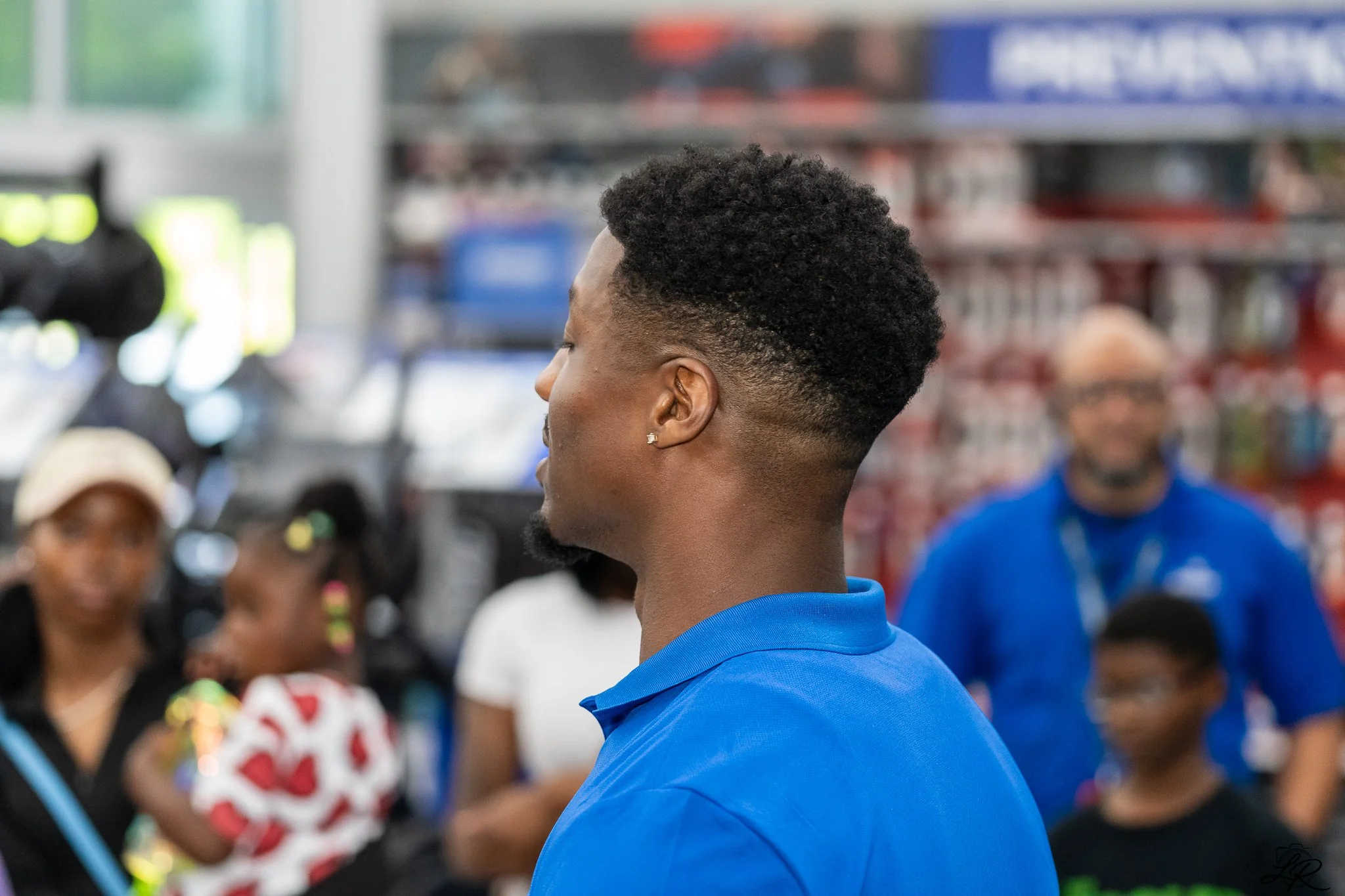 Profile of a young man with short, curly hair in a store, wearing a blue shirt, with a blurred background of people and store shelves.