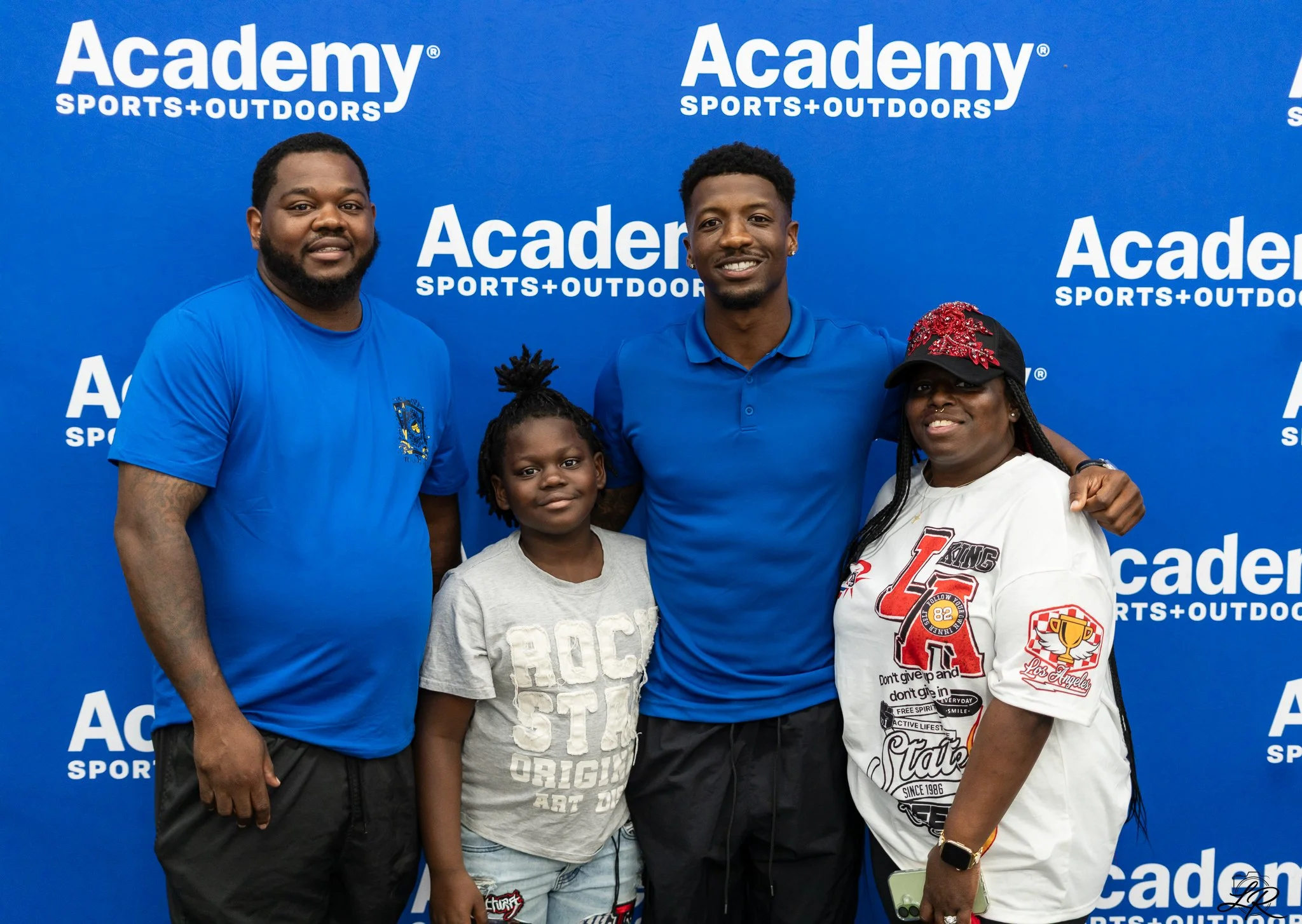 A group of five people, including two adults and three children, posing in front of a blue backdrop with white text that reads "Academy Sports + Outdoors" repeatedly. The group is smiling and dressed casually.
