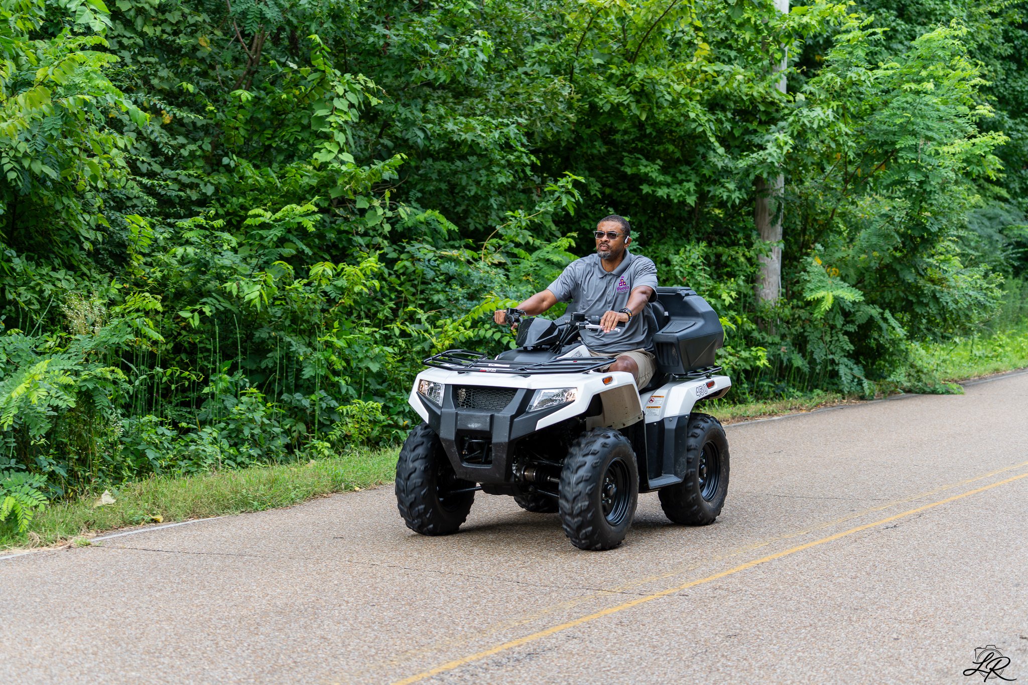 A man riding an all-terrain vehicle (ATV) on a paved road next to dense green trees and bushes.
