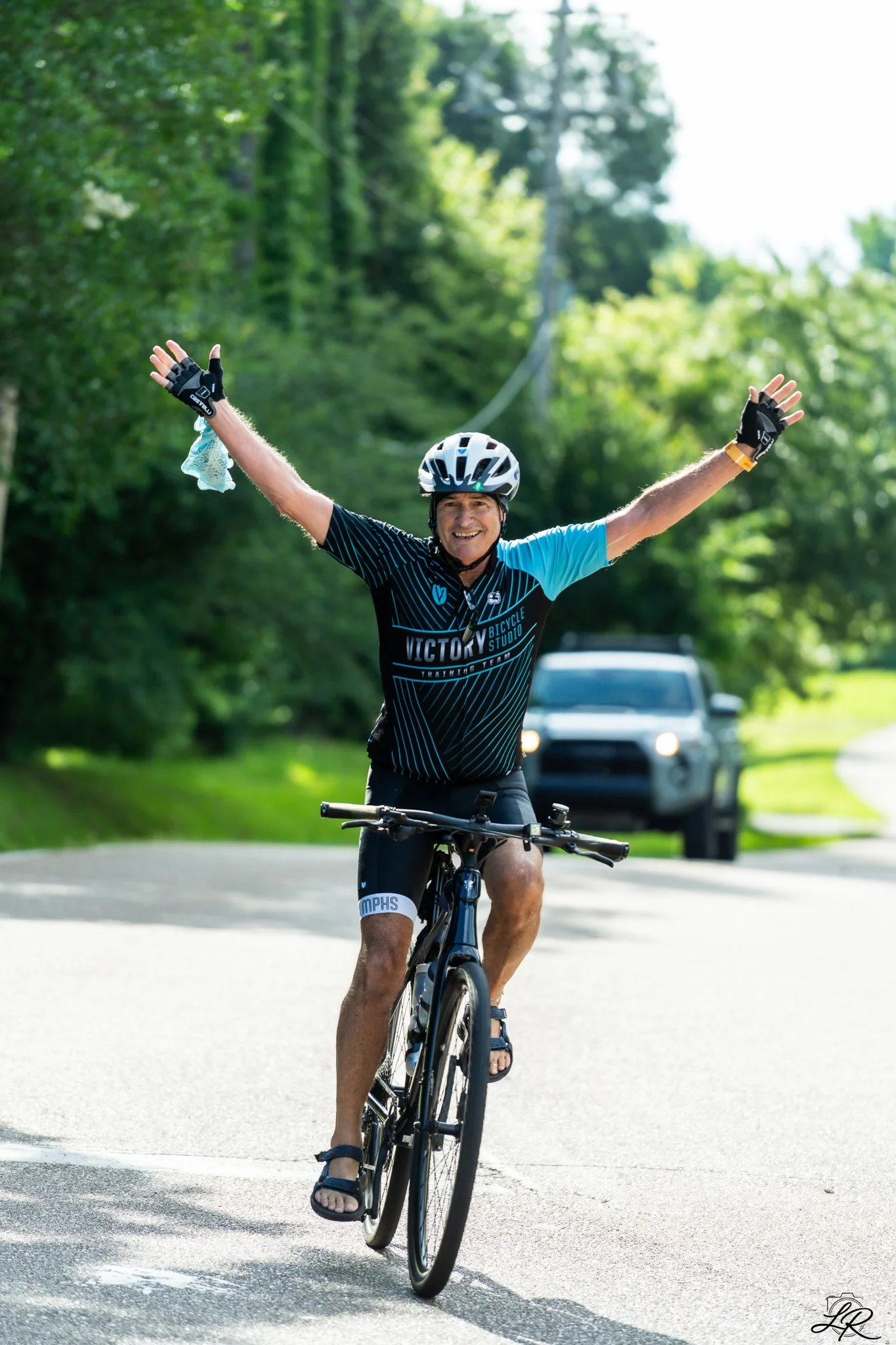 A man riding a mountain bike on a paved road with green trees in the background. He is wearing a helmet, cycling gloves, and a black and blue cycling jersey with the text 'Victory Bicycle Studio.' The man has his arms raised in celebration and is smi