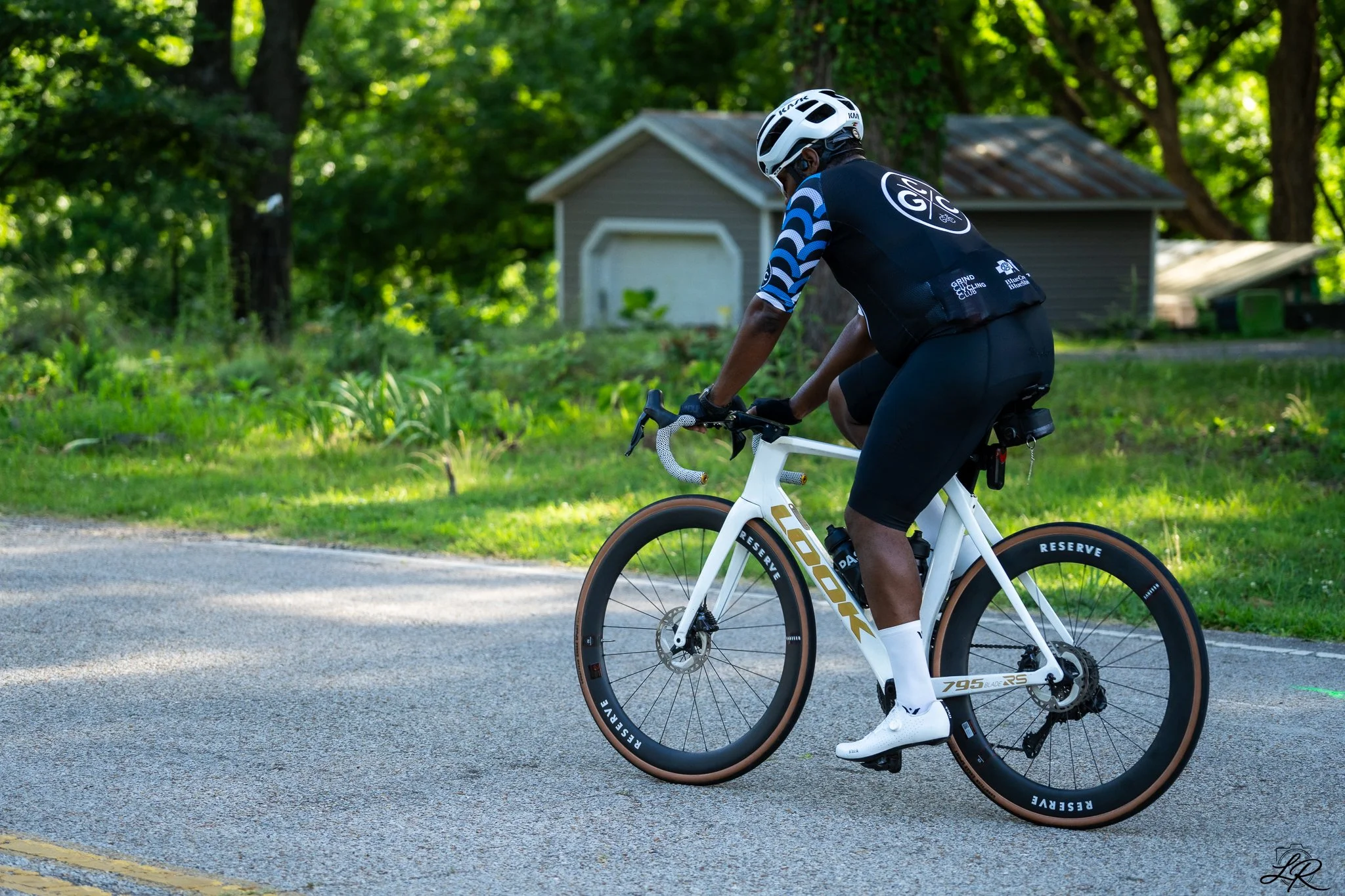 A person riding a white bicycle on a road surrounded by lush green trees, wearing a black helmet and cycling attire.