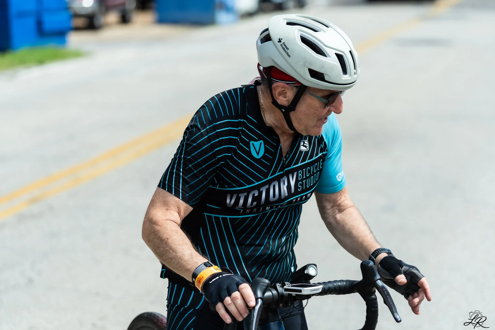 Older man riding a bicycle, wearing a white helmet, sunglasses, and a black cycling jersey with blue and white design.