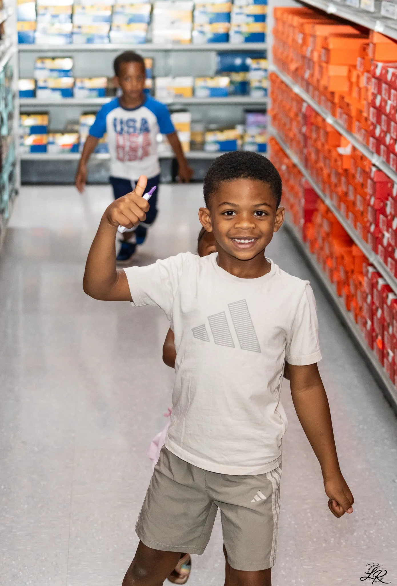 A smiling young boy in a white t-shirt and beige shorts giving a thumbs up while shopping in a grocery store aisle.
