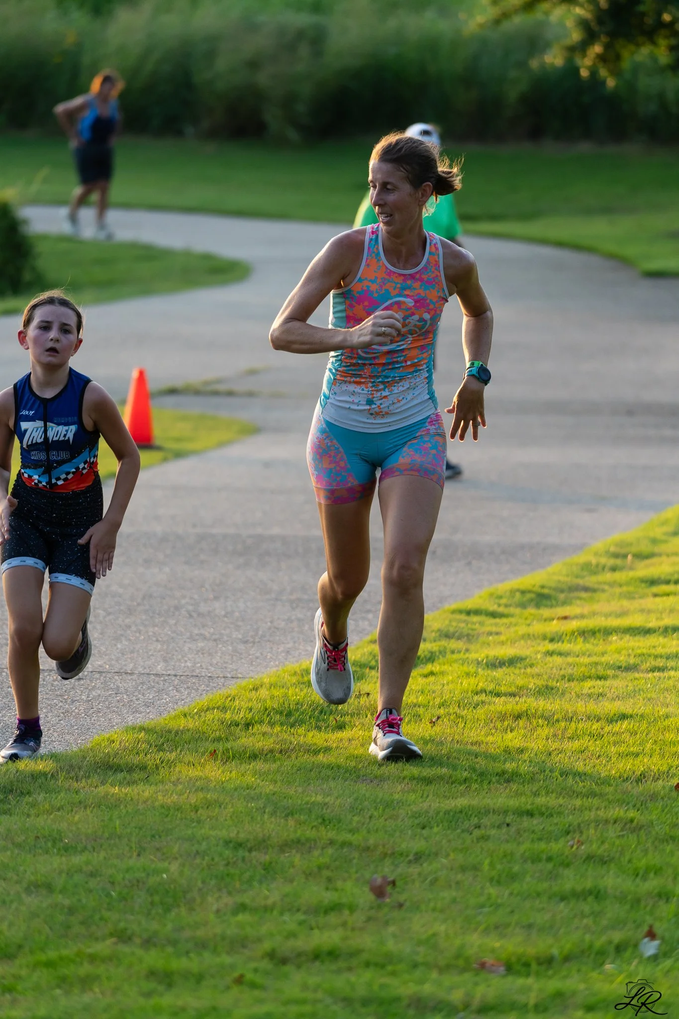 A woman and a young girl running outdoors on a paved trail through a grassy area at sunset, with other people in the background.