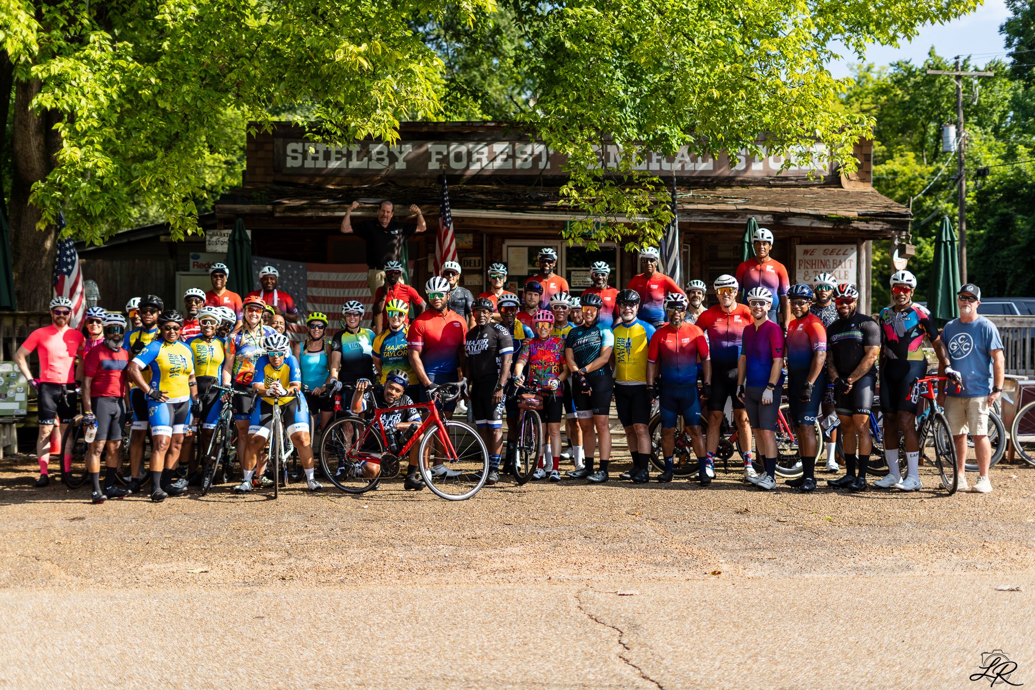 Group of cyclists and a man with arms raised in front of a rustic wooden building labeled Shelby Forest Bait & Tackle, surrounded by trees and American flags.